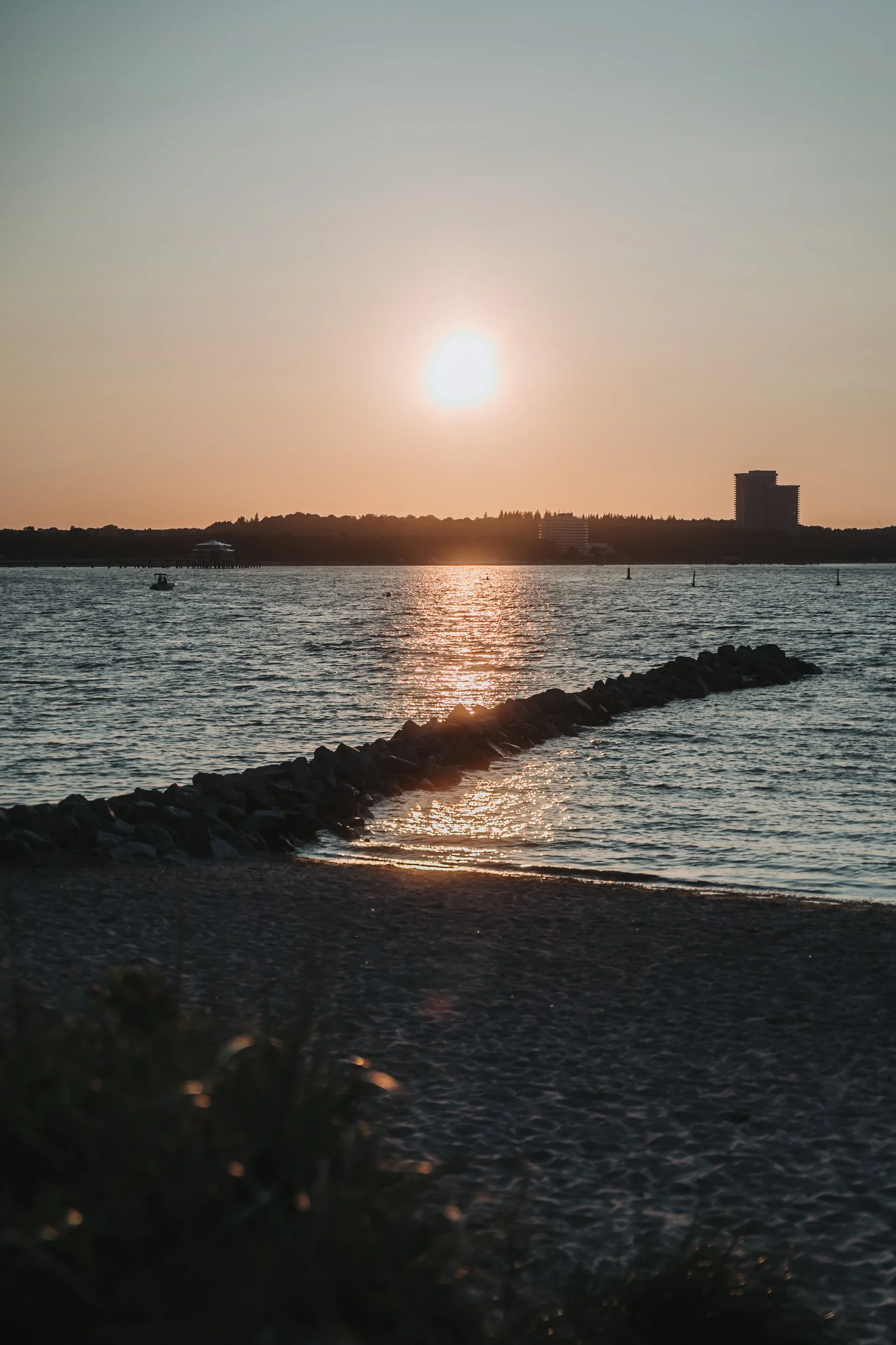 Sunset over a body of water with a silhouette of a shoreline, a boat on the water, and buildings in the distance.