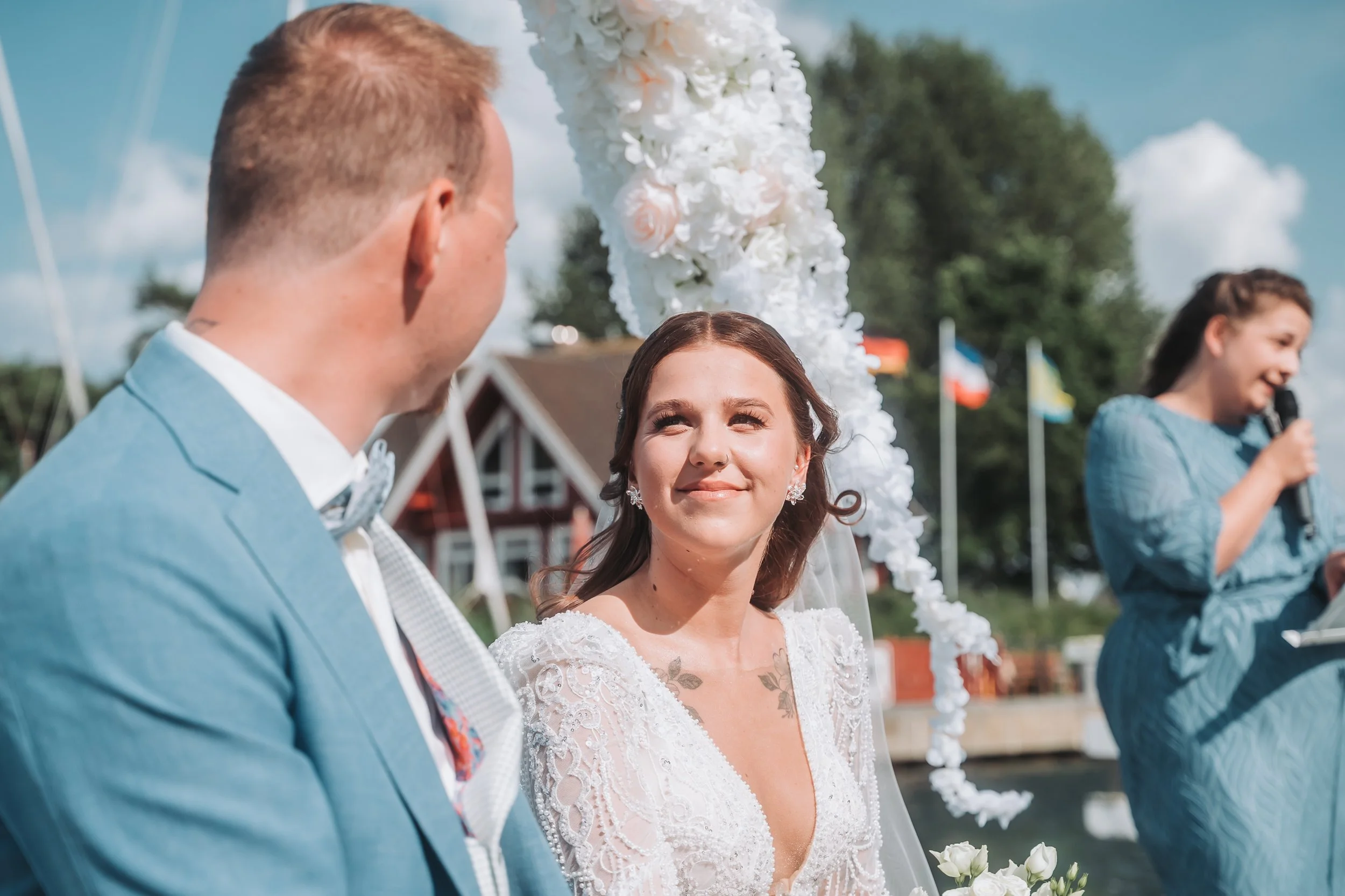 Bride with dark hair and earrings smiling while looking at groom, in a wedding ceremony outdoors on a sunny day, with a floral arch and flags in the background, and a bridesmaid holding a microphone.