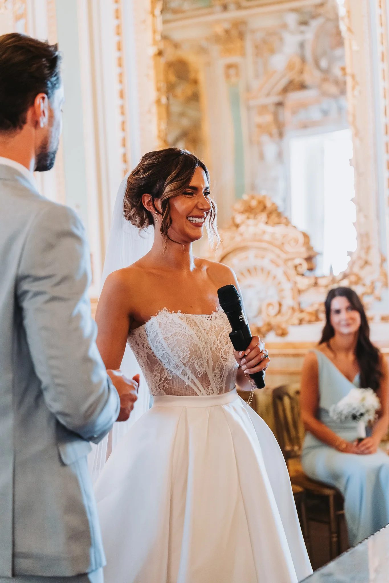 Bride smiling and holding a microphone during a wedding ceremony in an ornate, gold-accented room with a man in a light-colored suit and seated women in the background.