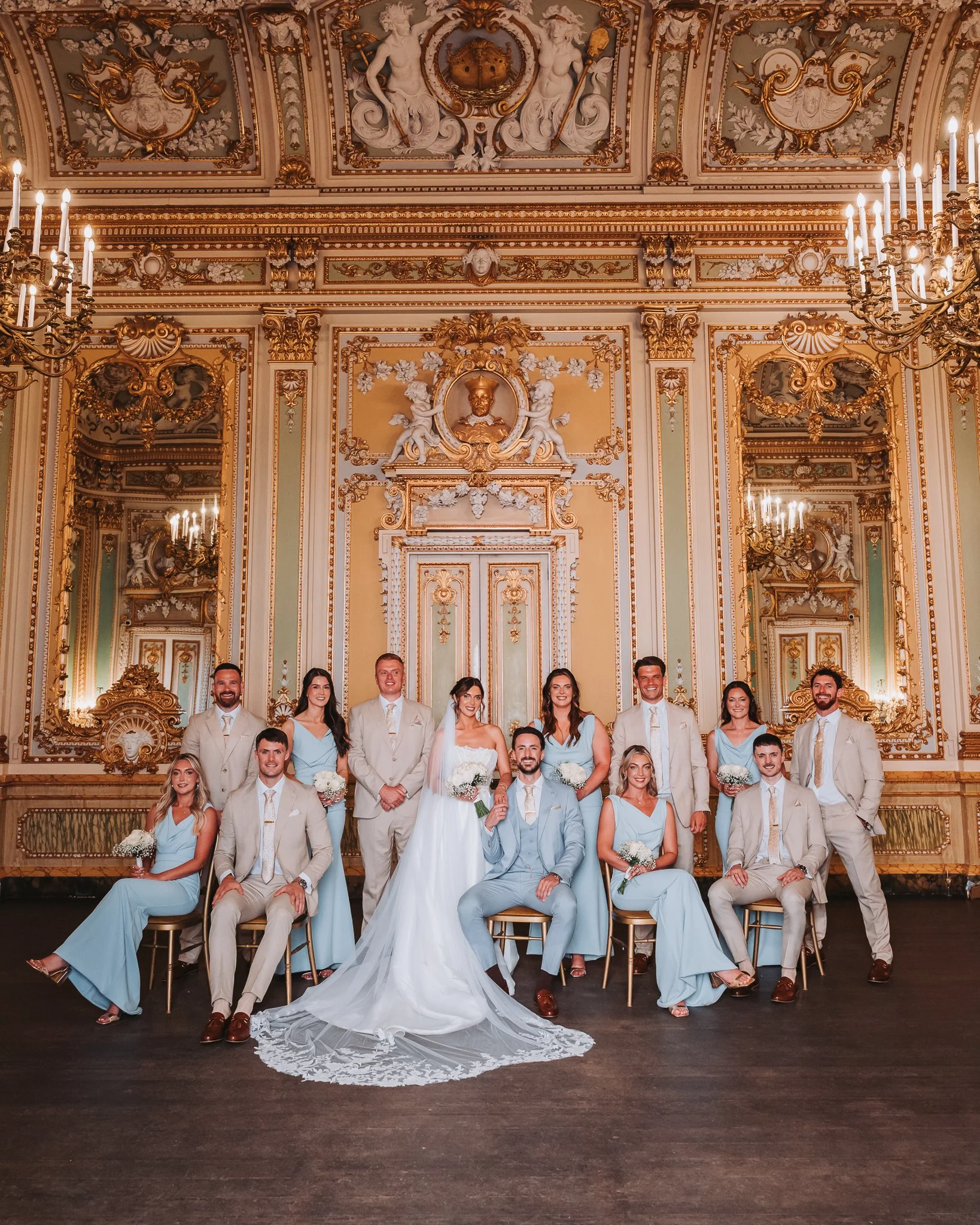 A wedding party posing in an ornate, gold-accented room with elaborate ceiling artwork. The group includes women in light blue dresses and men in beige suits. The bride in a white wedding gown and the groom in a light blue suit sit in the center, sur