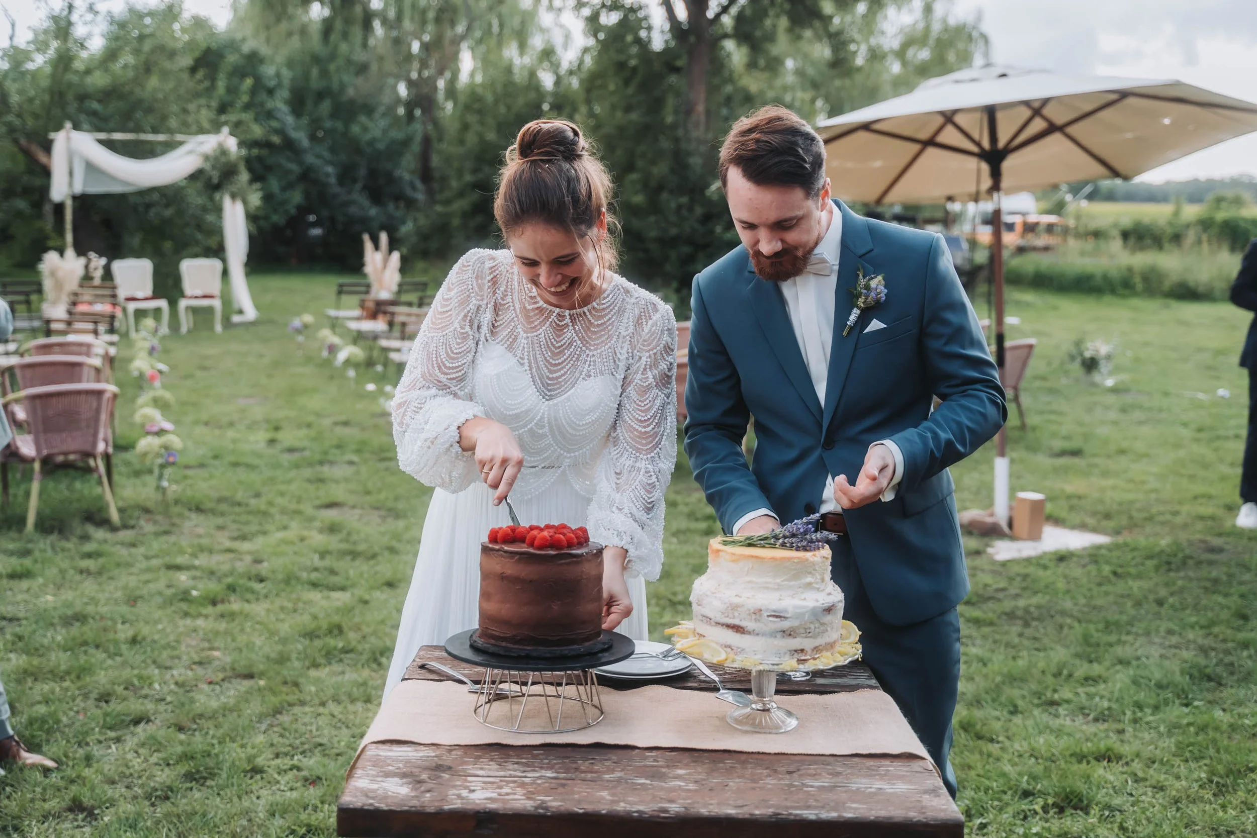 A bride and groom cutting wedding cakes outdoors during their wedding reception with chairs, tables, and umbrellas in the background.