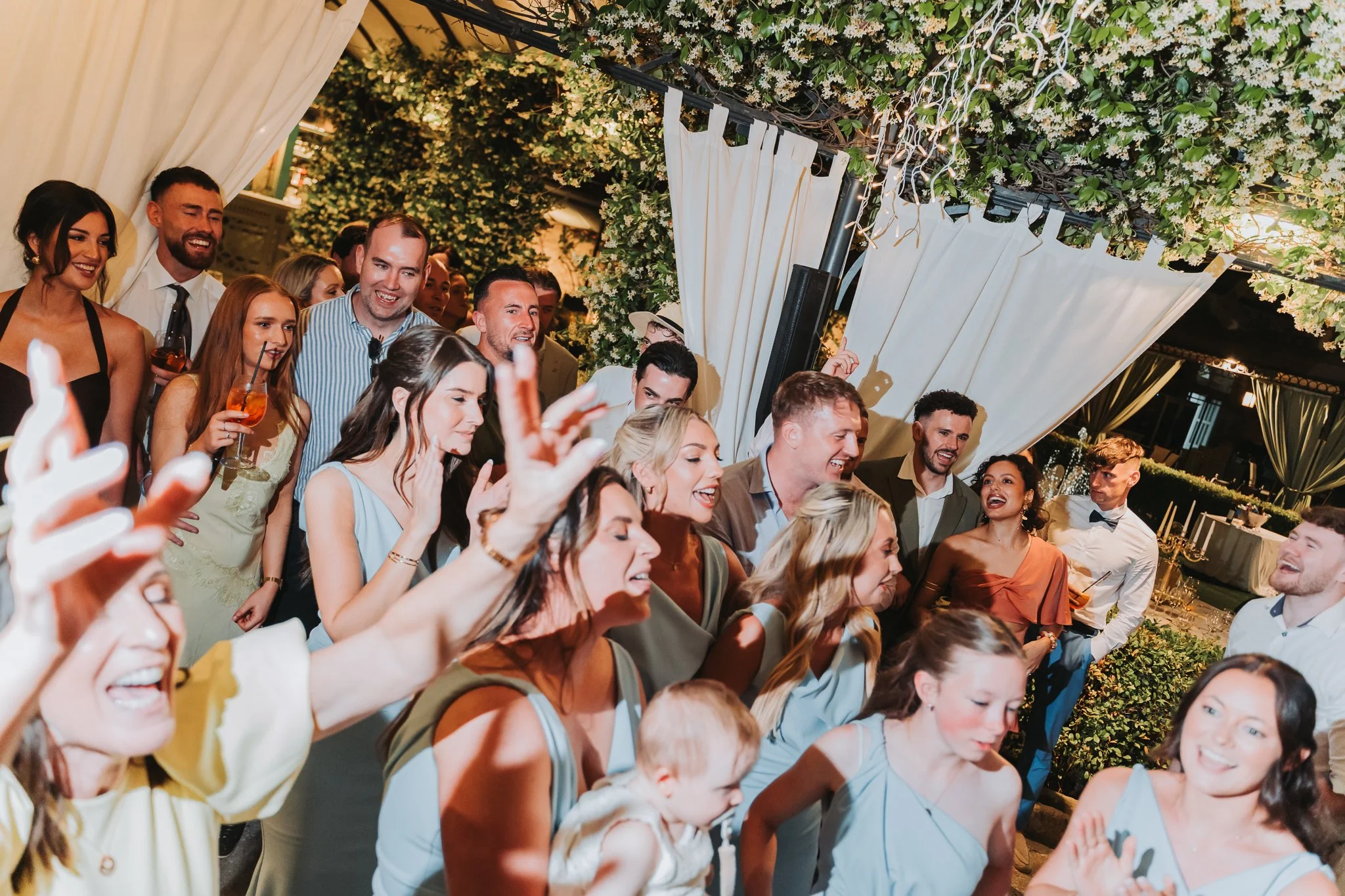 Group of people at a celebration or party, smiling and enjoying the moment, decorated with floral arrangements and drapery.