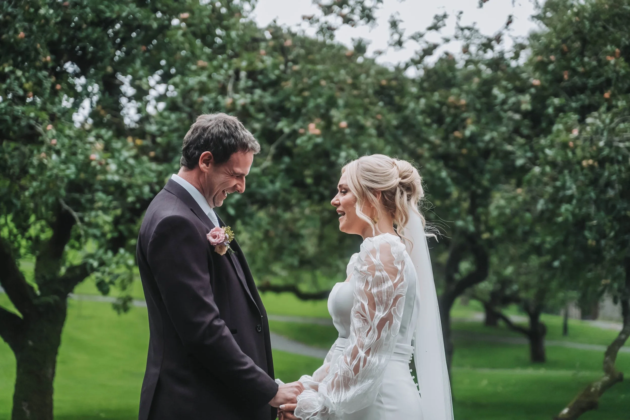 A bride and groom holding hands and smiling at each other outdoors surrounded by green trees during a wedding ceremony.