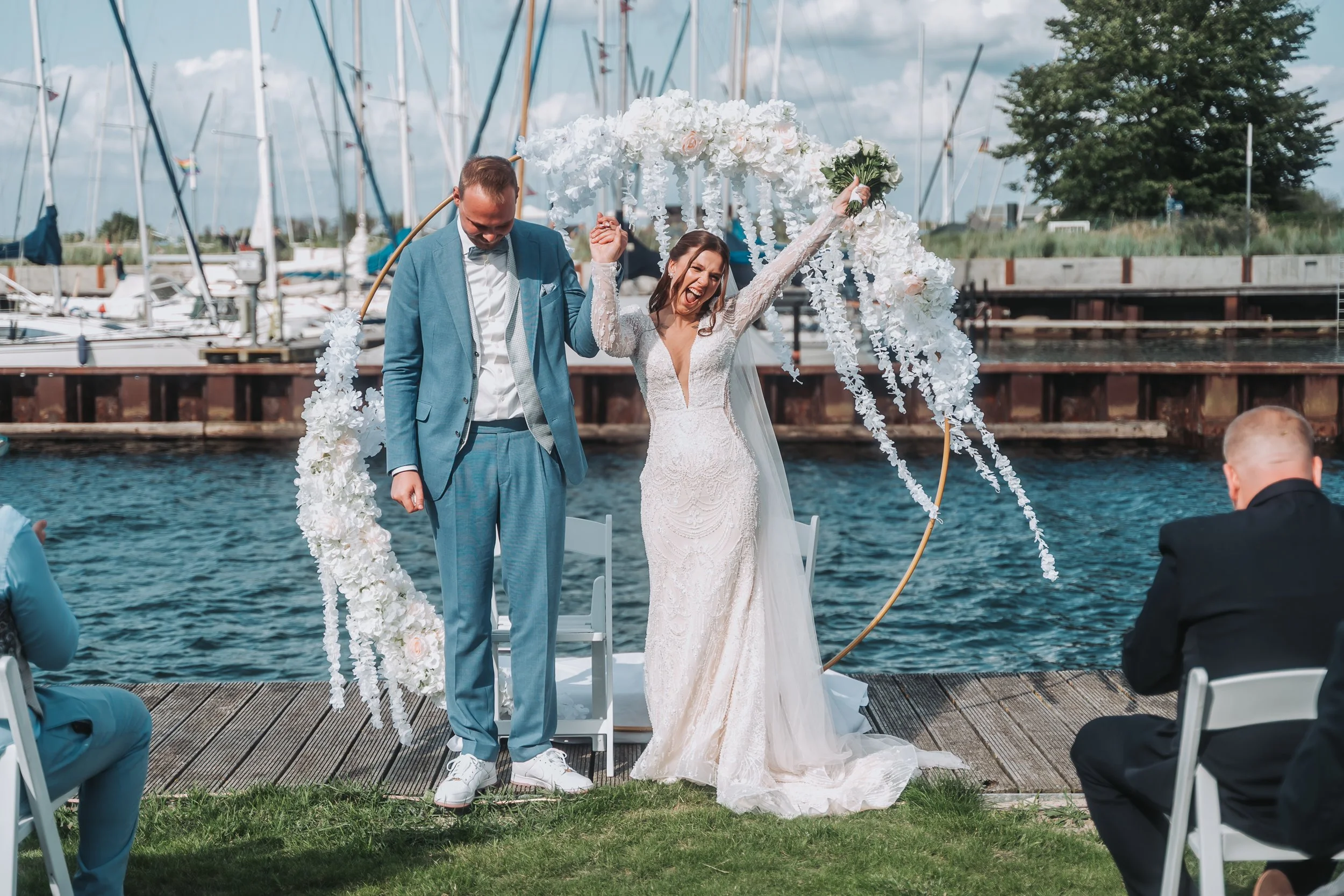 A couple getting married outdoors by a marina, with the bride holding a bouquet and raising her arm, and the groom standing next to her, both smiling.