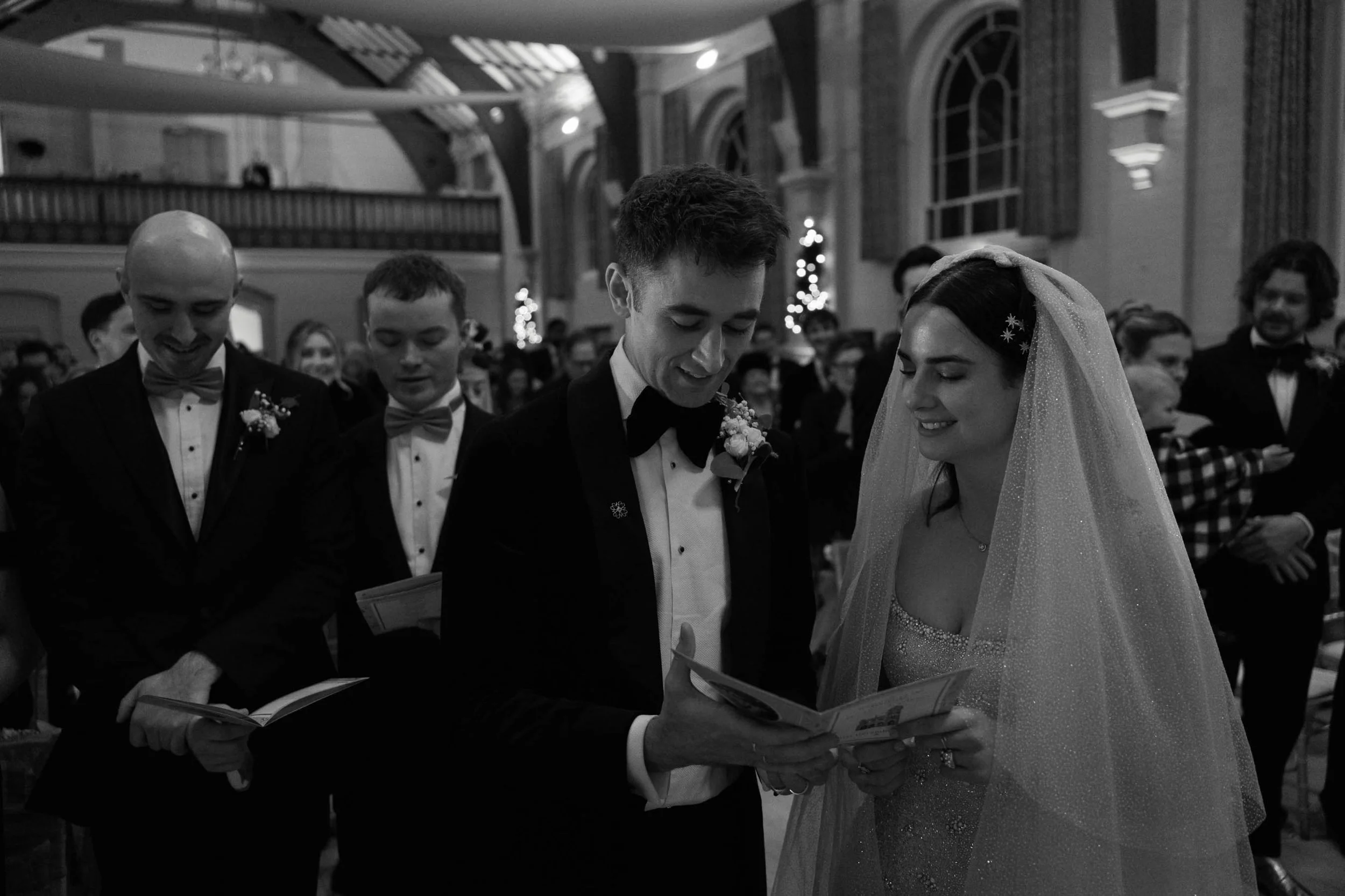A black and white photo of a wedding ceremony inside a church with a group of people, including a bride and groom, standing close together and reading from a booklet. The bride is wearing a veil and a sparkly dress, while the groom is dressed in a tu