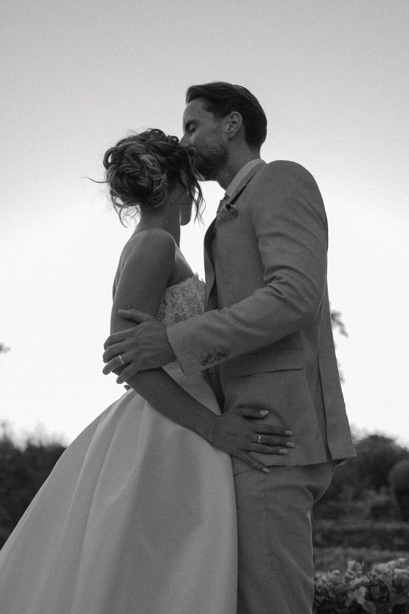A black and white photo of a bride and groom embracing outdoors at sunset, with the bride wearing a strapless lace wedding gown and the groom in a suit, sharing a tender moment.