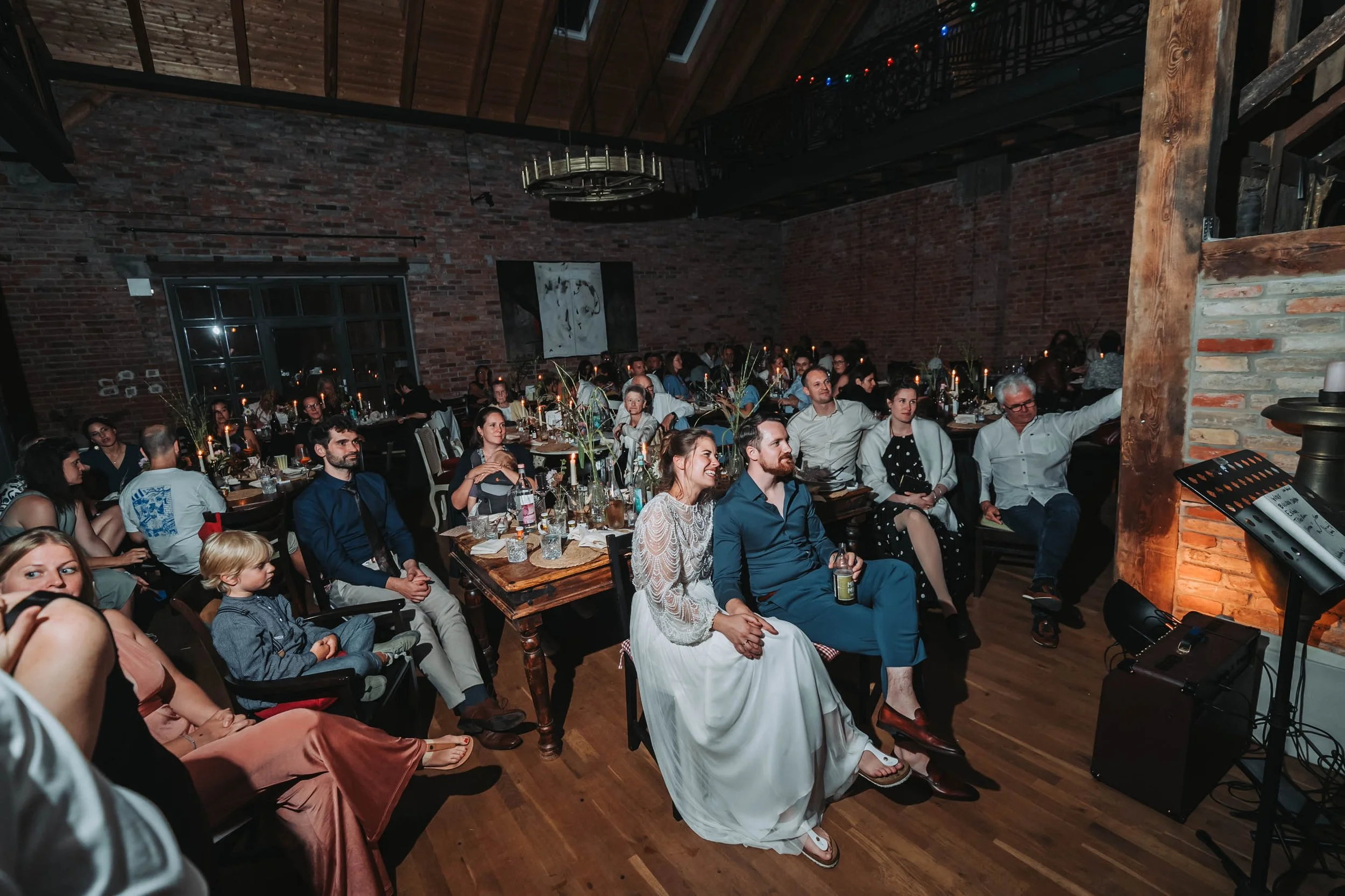 People sitting and standing in a dimly lit, rustic restaurant with exposed brick walls and wooden beams, watching a performance or presentation.
