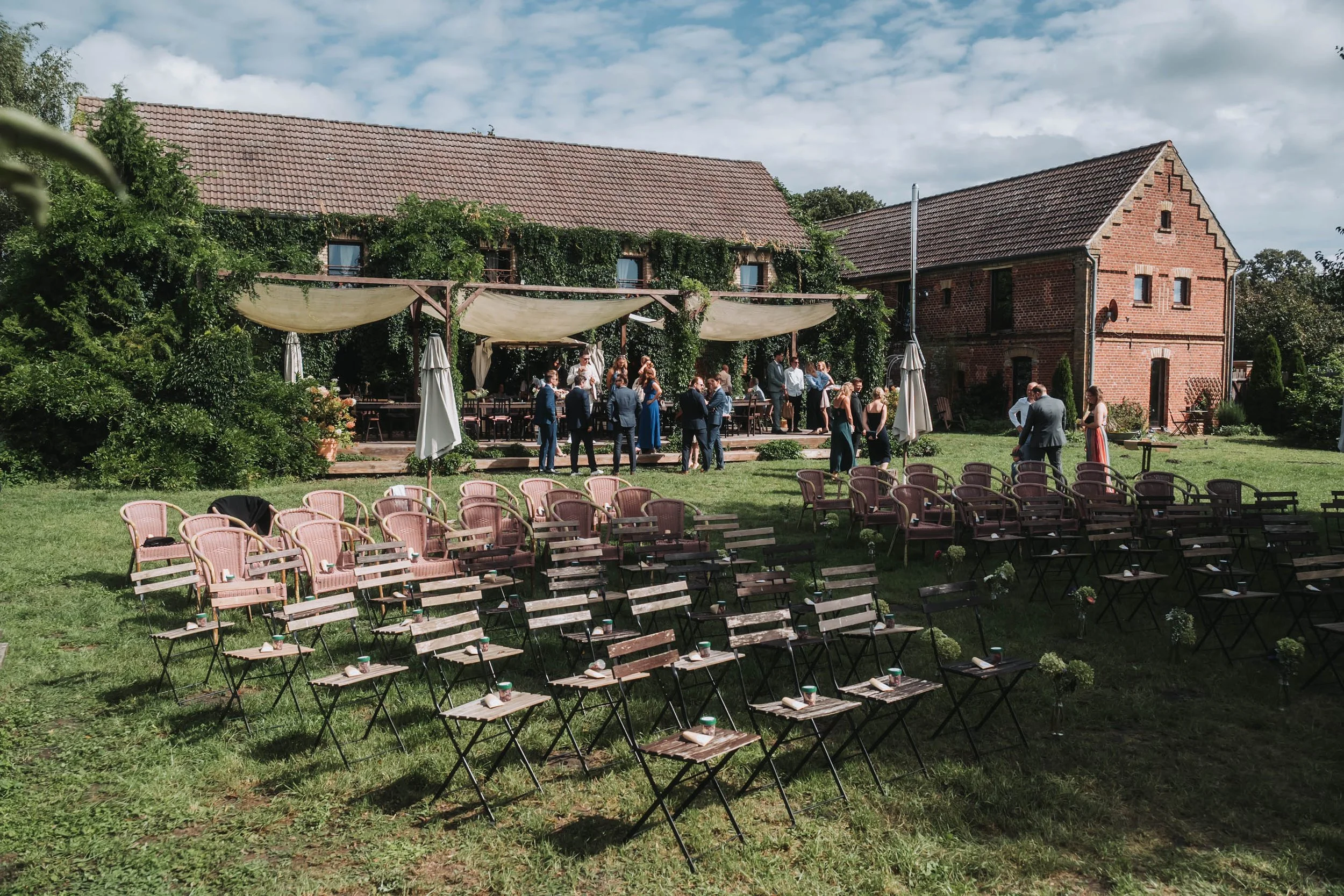 Outdoor gathering with people dressed in formal attire, standing and sitting on pink chairs in a grassy area, with brick buildings covered in green vines and a partially cloudy sky in the background.