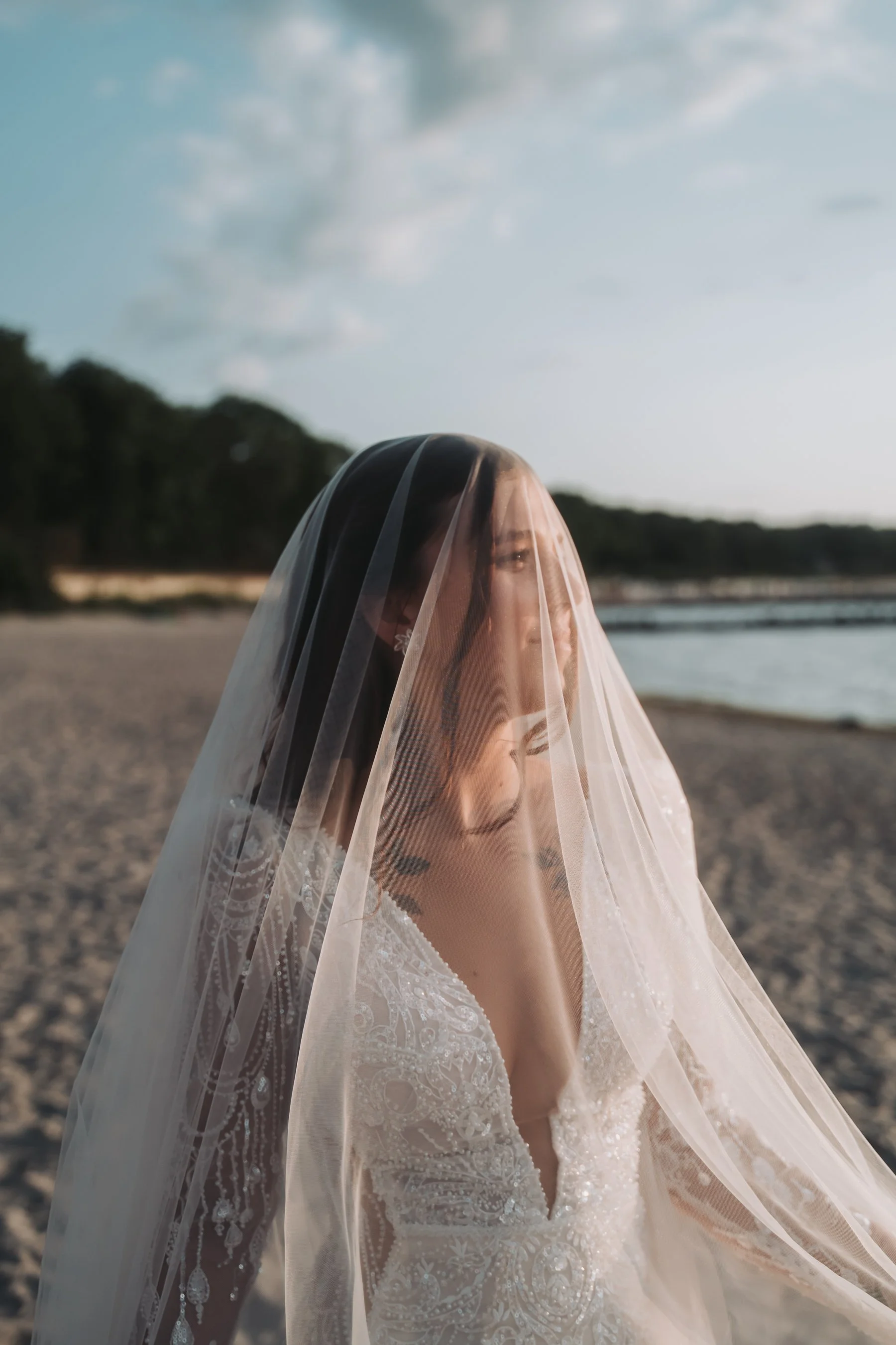 A woman wearing a white lace dress and a sheer veil standing on a sandy beach by the ocean, with trees and a partly cloudy sky in the background.