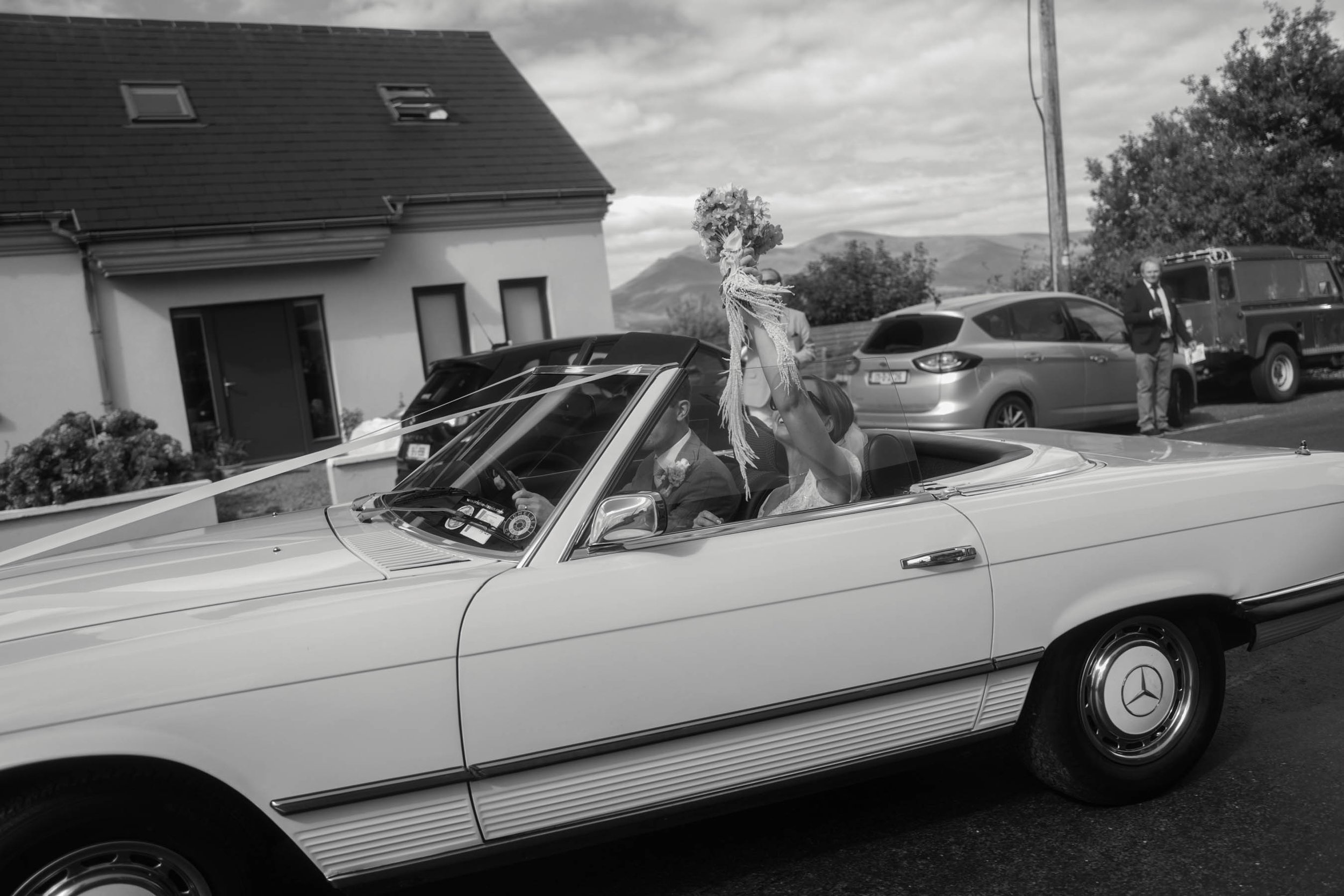A vintage convertible car with a bride and groom inside, the bride holding a bouquet of flowers. The car is decorated for a wedding. There are people and parked cars in the background, with a house and mountains in the distance.