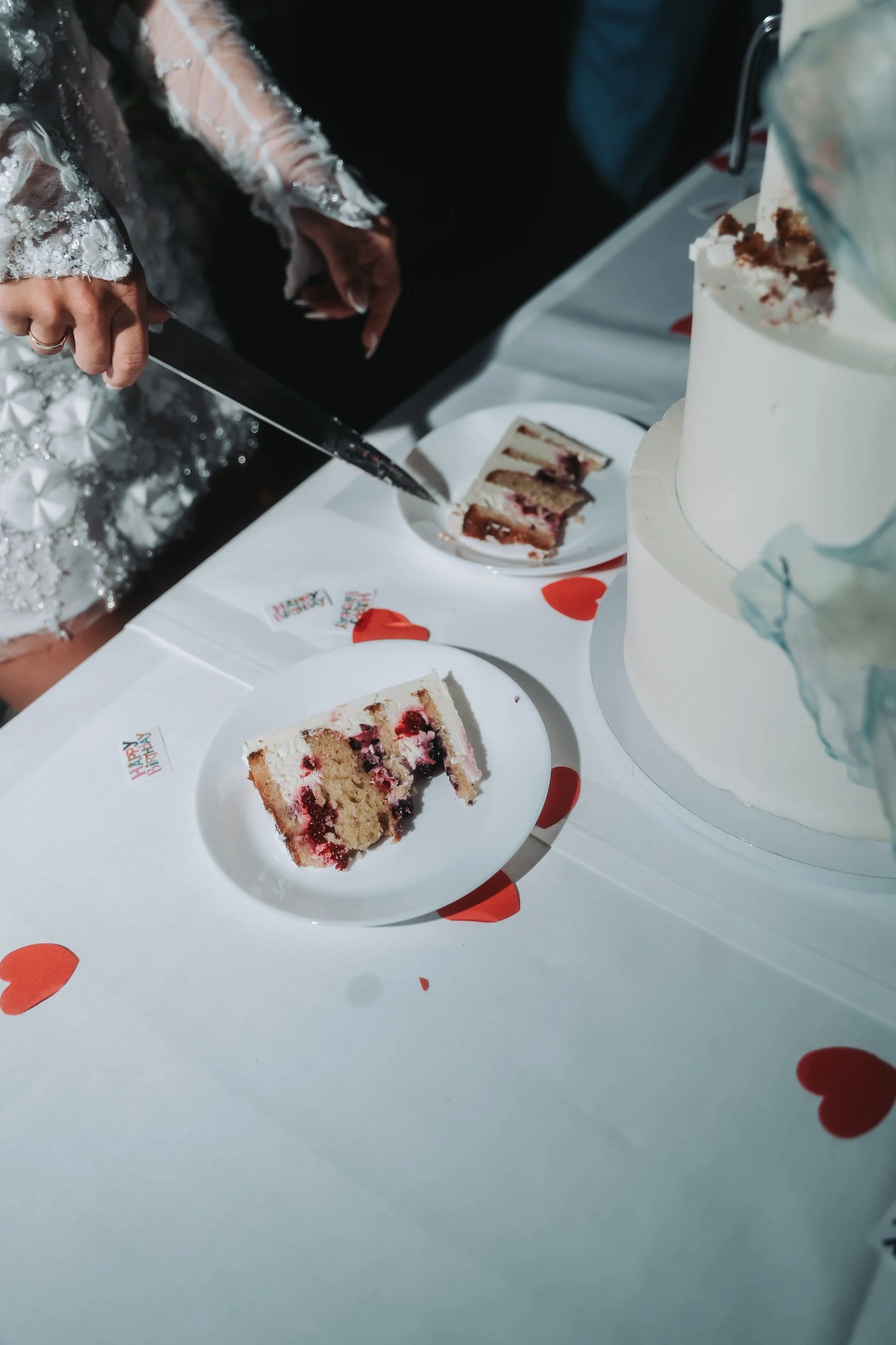 A person in a lace and beaded dress is cutting a slice of cake with a knife. The cake has layers of white frosting, red fruit filling, and cake layers. The table is decorated with small 