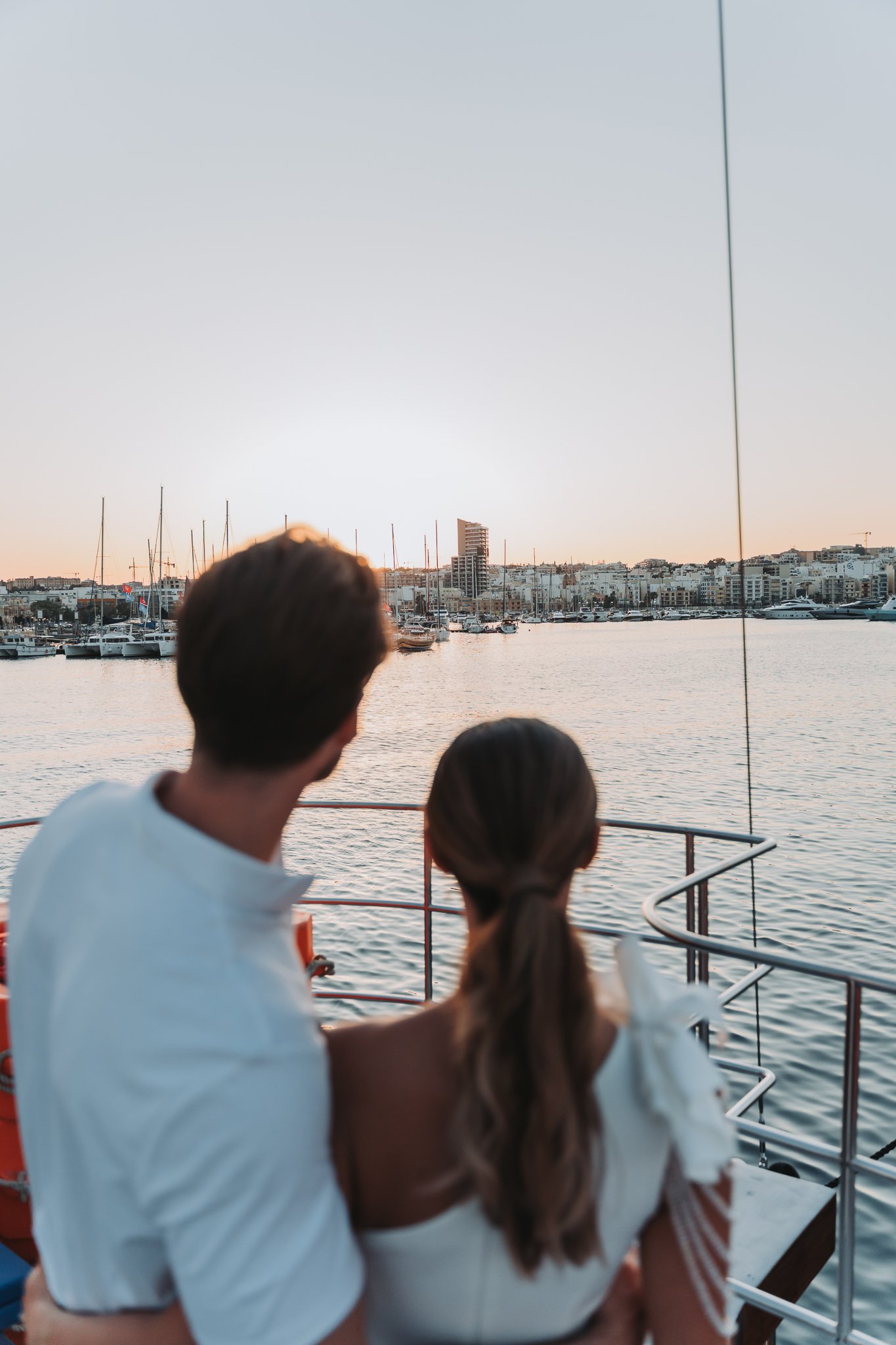A couple standing on a boat deck, with water and a city skyline with boats and yachts in the background, during sunset.