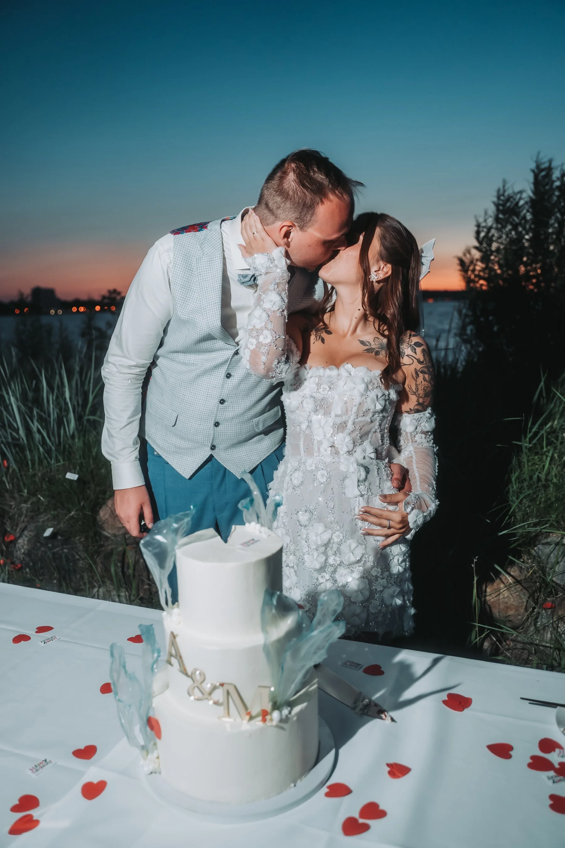 A newlywed couple sharing a kiss during their outdoor wedding reception at sunset. The bride wears a white lace wedding dress, and the groom wears a light-colored vest and shirt. There is a white wedding cake with gold letters on a table decorated wi
