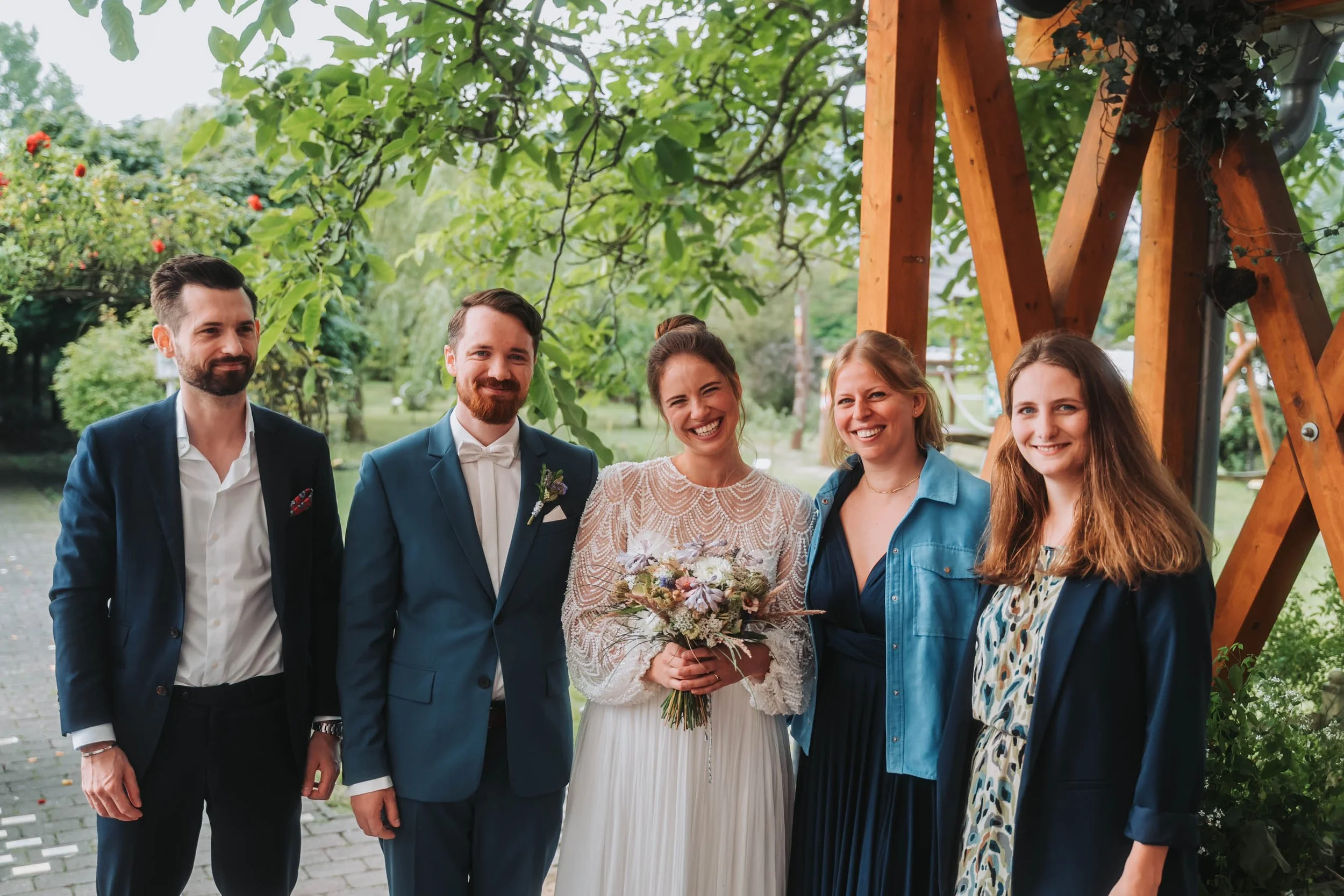 Group of five people standing outdoors, smiling, with a woman holding a bouquet of flowers in the middle. The group includes men and women dressed in semi-formal and casual attire, with lush green trees and a wooden structure in the background.