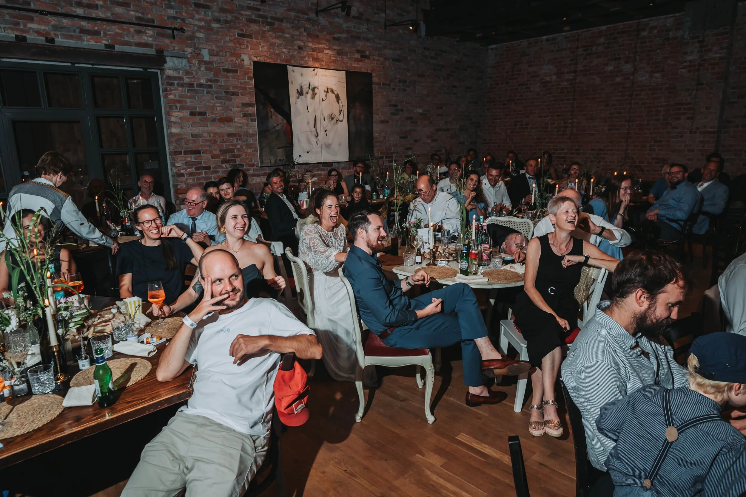 People sitting at tables in a lively indoor event or celebration with exposed brick walls and abstract artwork on the wall, many are smiling and laughing.