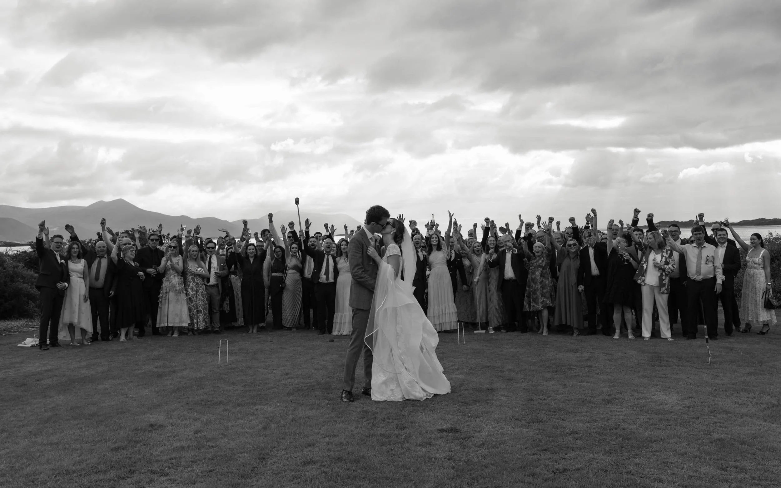 A wedding celebration outdoors with many guests, a bride and groom in the foreground kissing, and mountains in the background under cloudy skies.