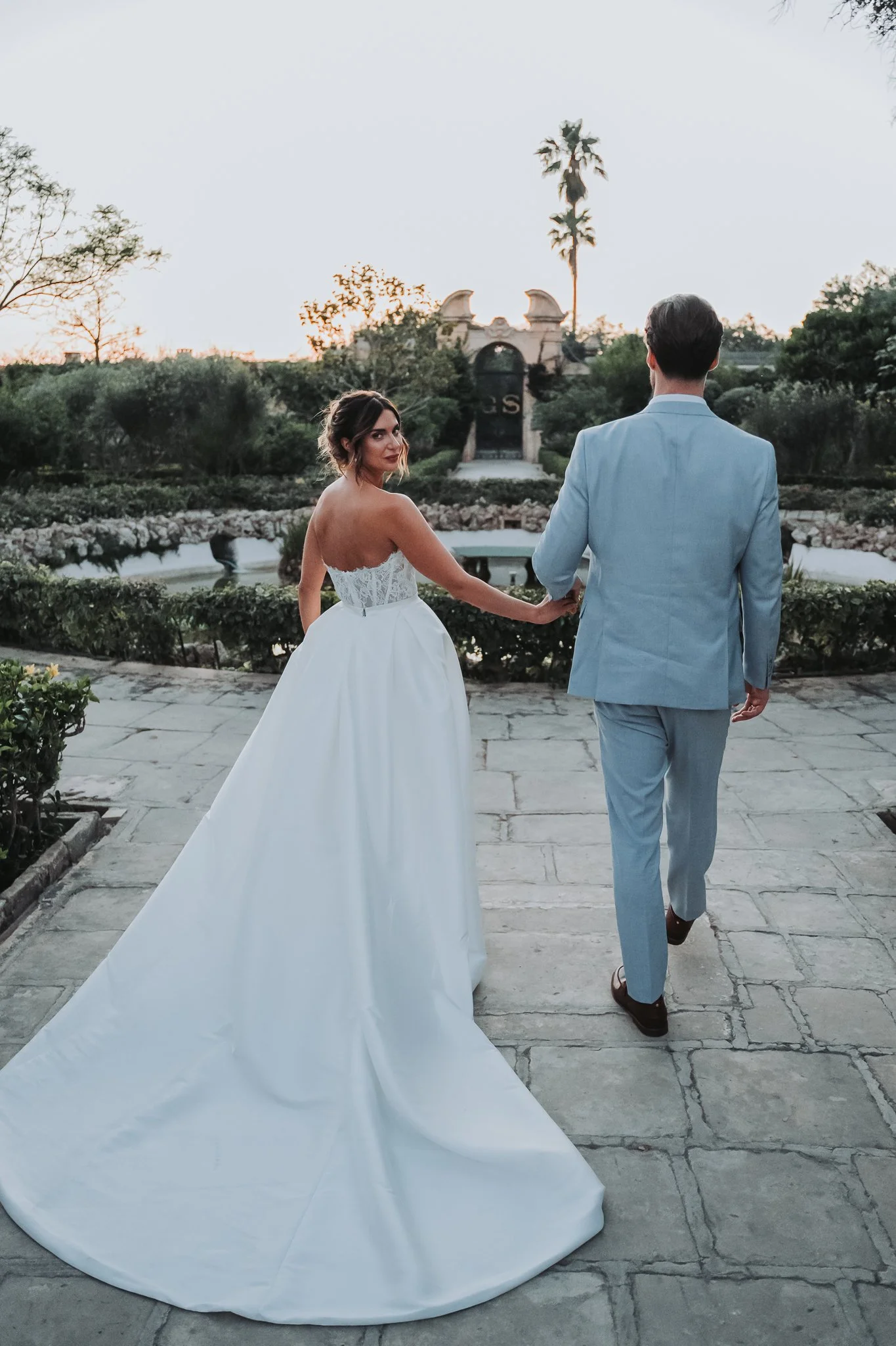 A bride and groom holding hands and walking outdoors at sunset, with a garden and historic archway in the background.