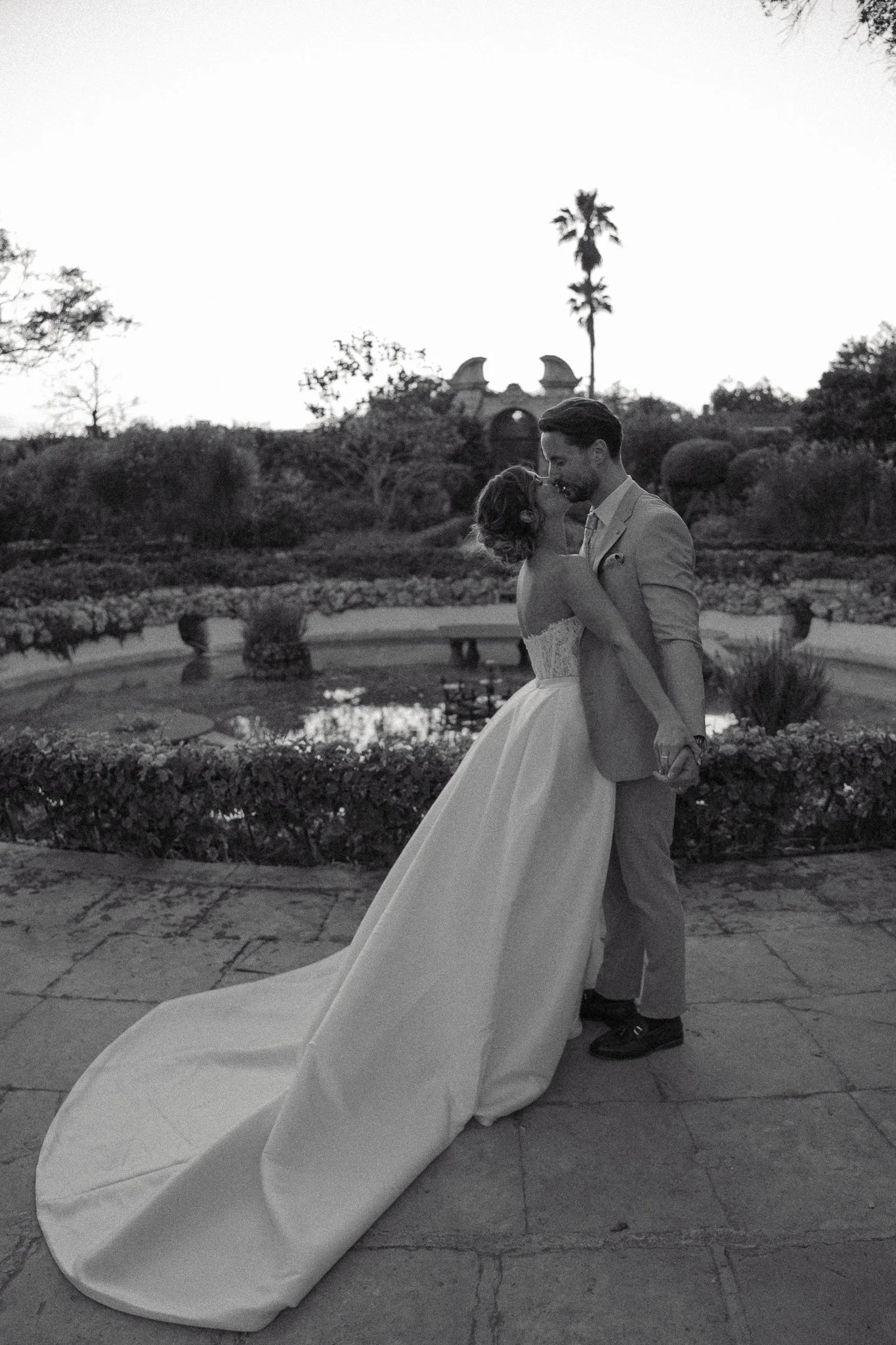 A couple sharing a romantic moment outdoors, with the woman in a wedding dress and the man in a suit, holding each other and about to kiss, in a garden with a pond and trees in the background.