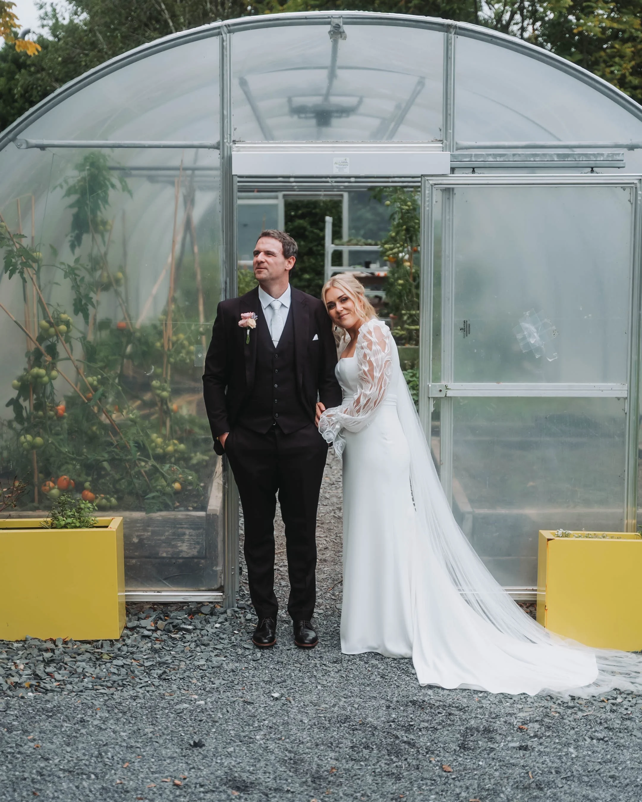 A man in a black suit and a woman in a white wedding dress stand together outside a glass greenhouse with tomato plants inside. It appears to be a wedding or special occasion.