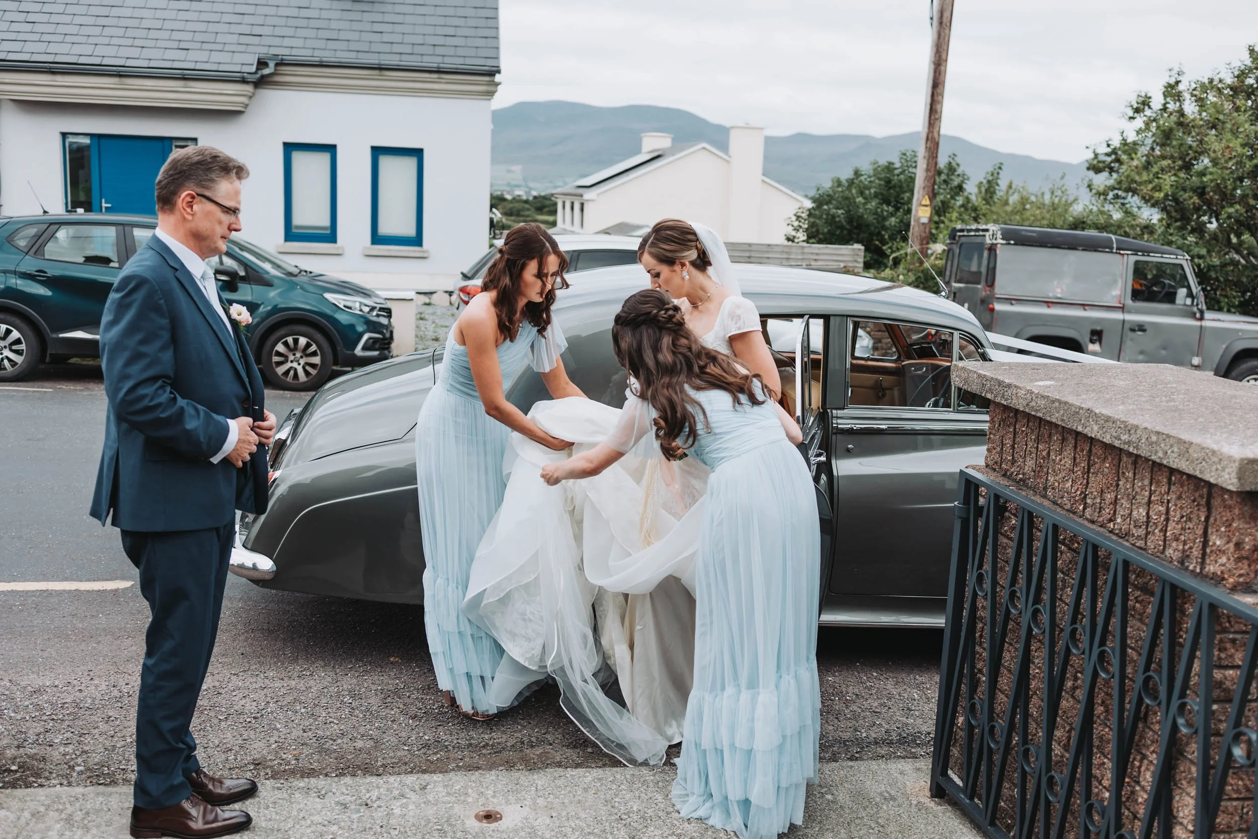 People dressed in formal attire, likely at a wedding, with wedding dresses and a man in a suit, standing next to a vintage black car on a street in a residential area with houses and mountains in the background.