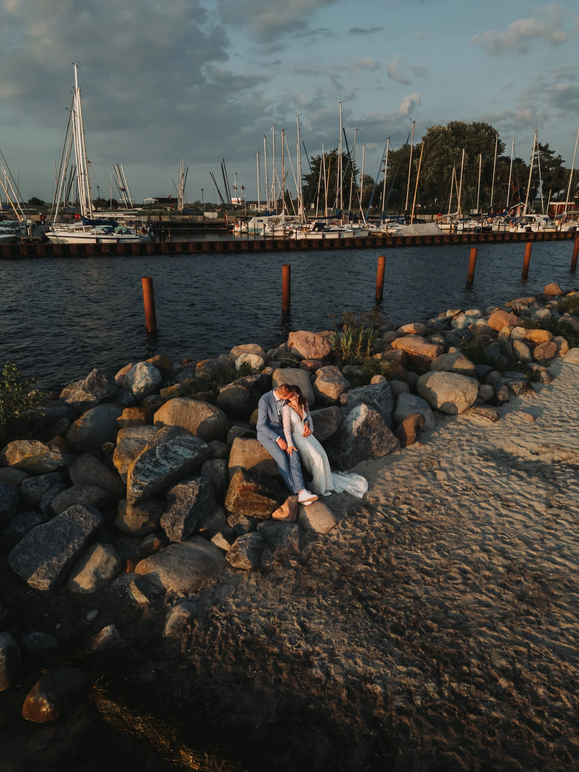 A couple sitting on a rocky shoreline at sunset by a marina with boats and sailboats docked in the water.