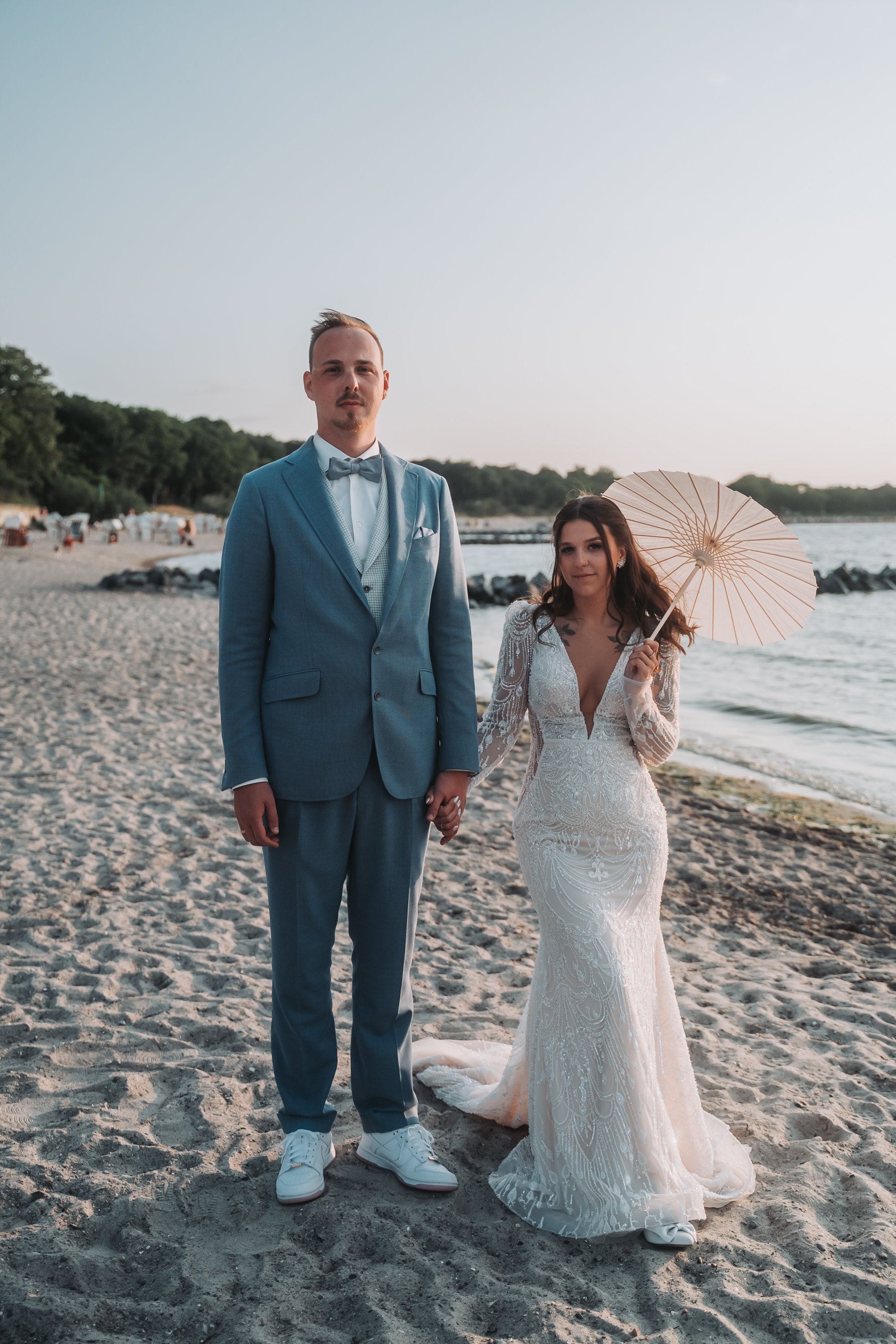 A bride and groom holding hands on a beach during sunset. The groom is wearing a blue suit with a bow tie, and the bride is wearing a white lace wedding gown and holding a pink parasol.