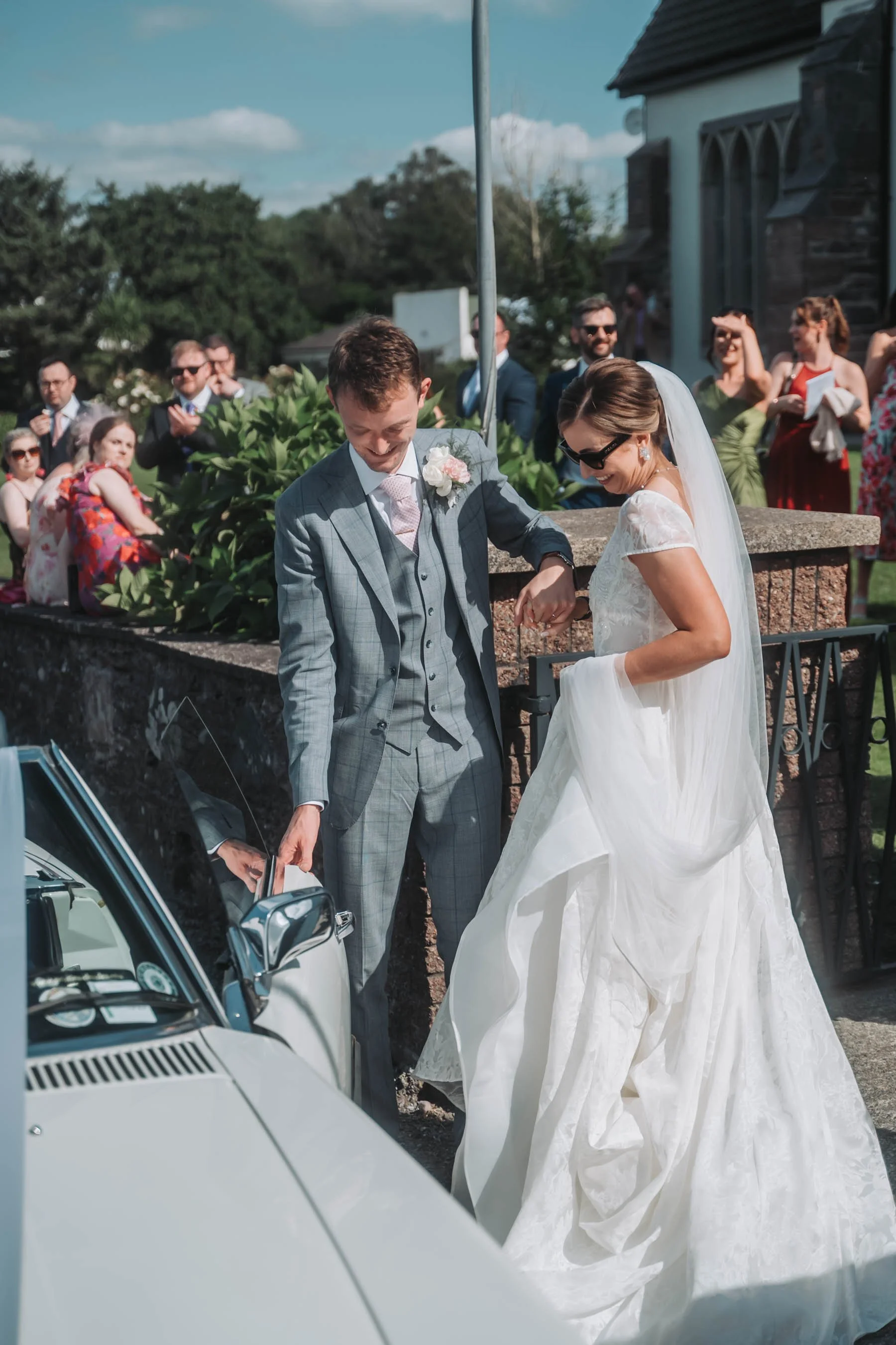 A bride and groom in wedding attire exchanging something from a car during a wedding celebration, surrounded by well-dressed guests.