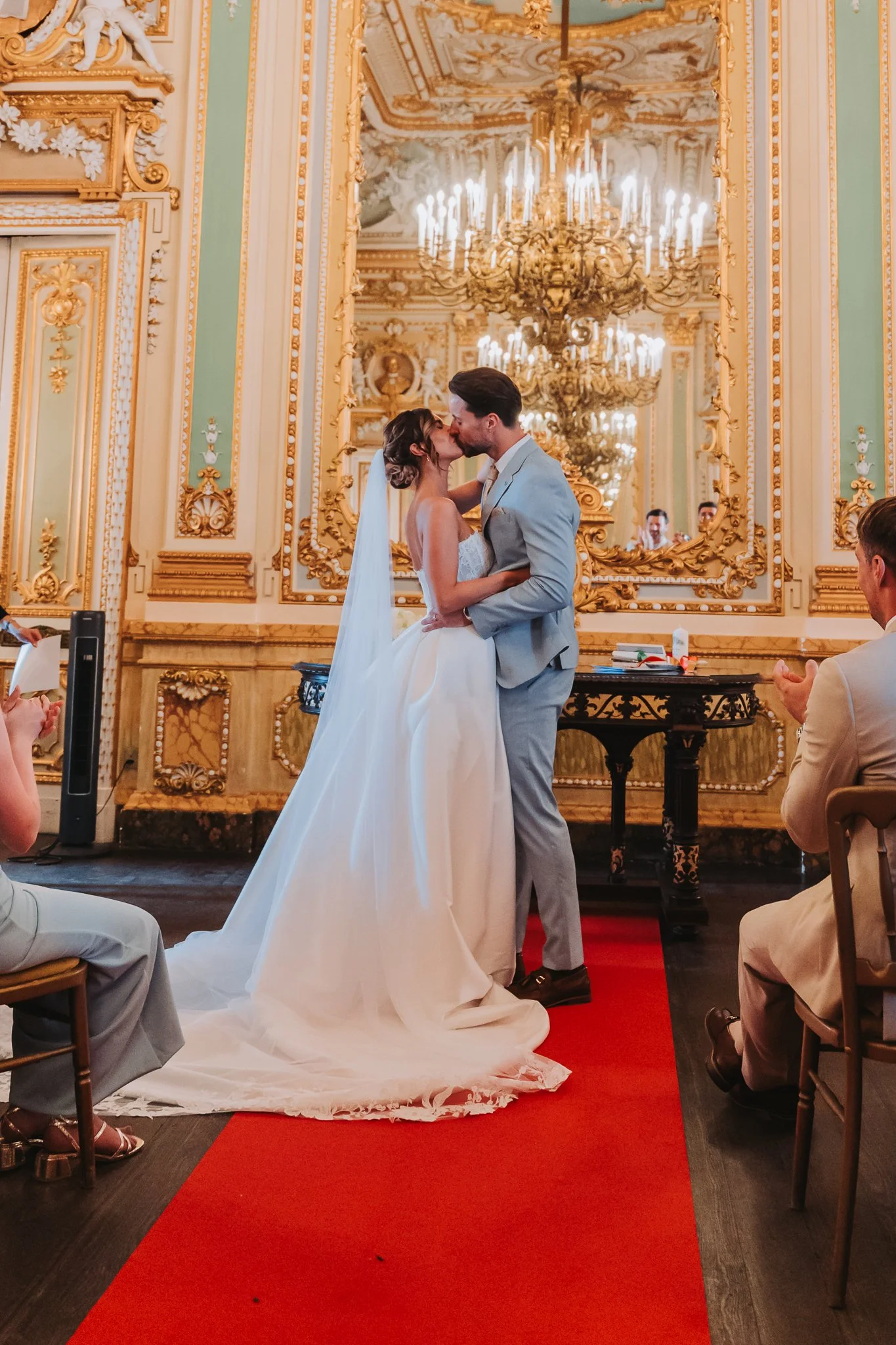 A couple is kissing during a wedding ceremony in an ornate, gold-adorned hall with chandeliers and a large mirror behind them.
