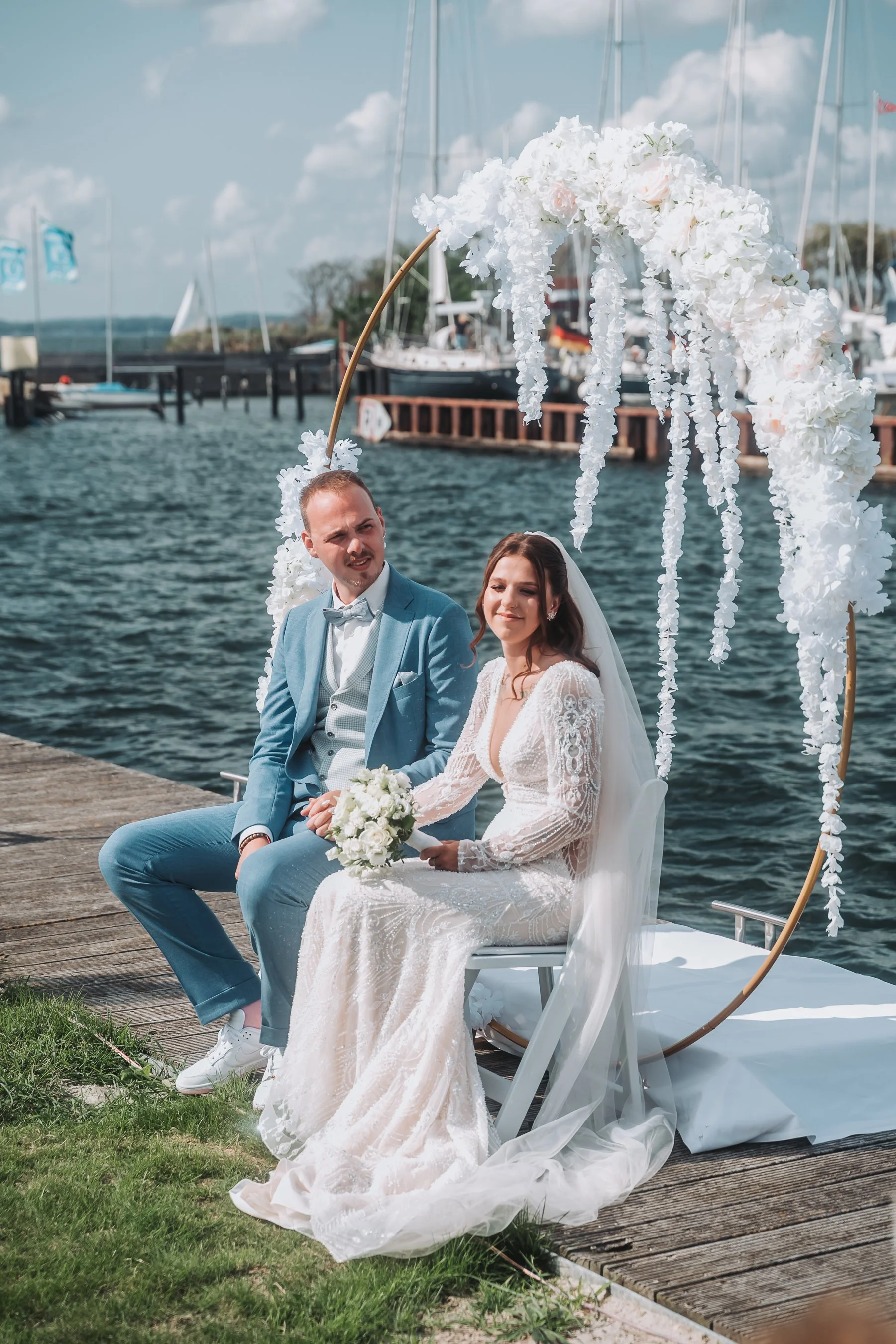 A newlywed couple sitting on a decorated boat docked by a lake or river during daytime. The groom is wearing a light blue suit with a bow tie, and the bride is in a white lace wedding dress holding a bouquet of white flowers. Behind them, white flora