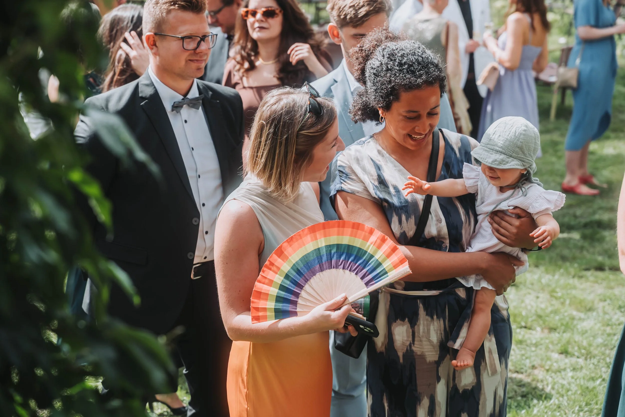 A group of people at an outdoor event, including women and men, some dressed in formal attire, holding a colorful rainbow fan, and engaging with a smiling baby wearing a hat.