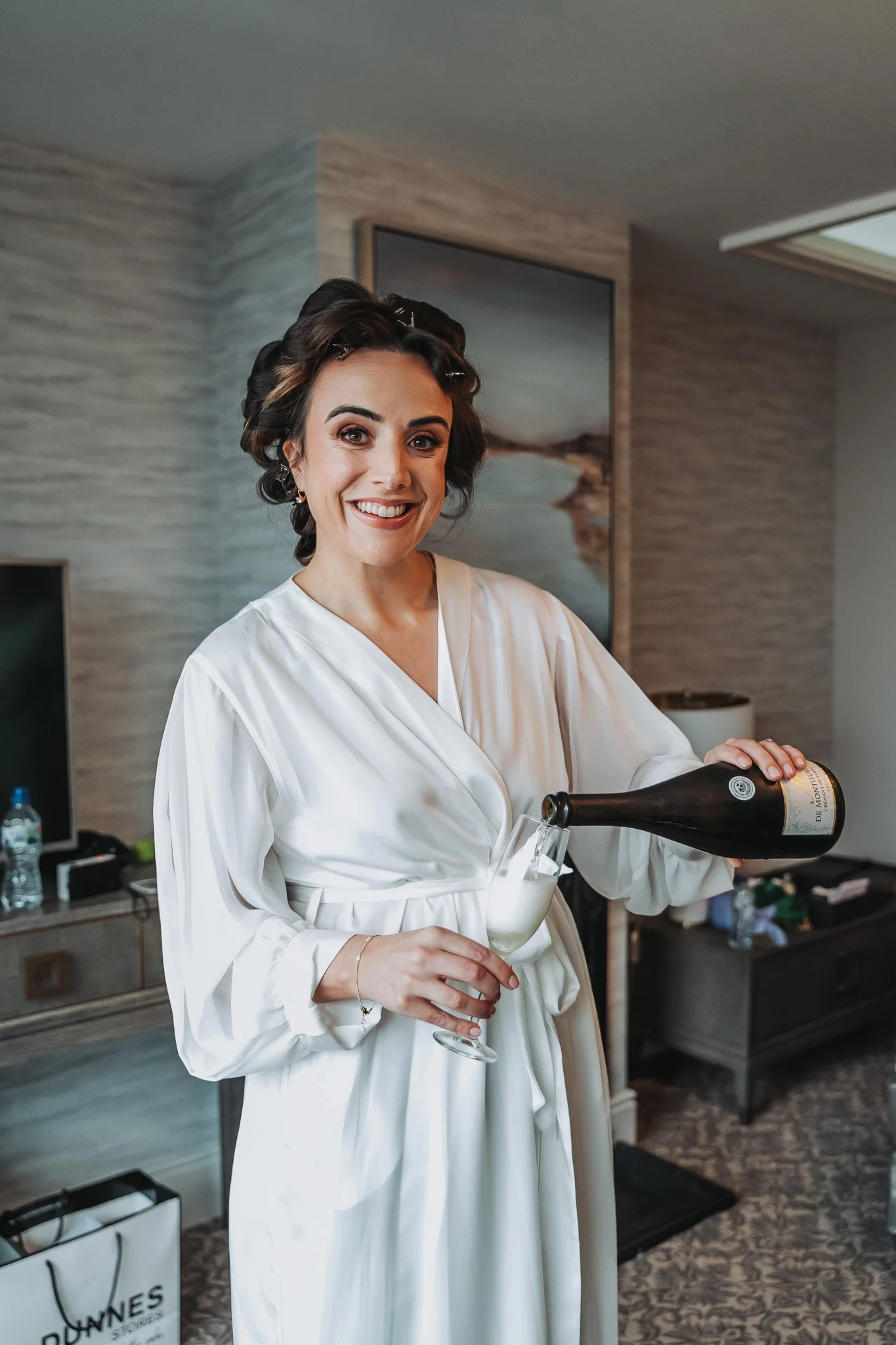A woman wearing a white robe and hair rollers, smiling, pouring champagne into a flute.