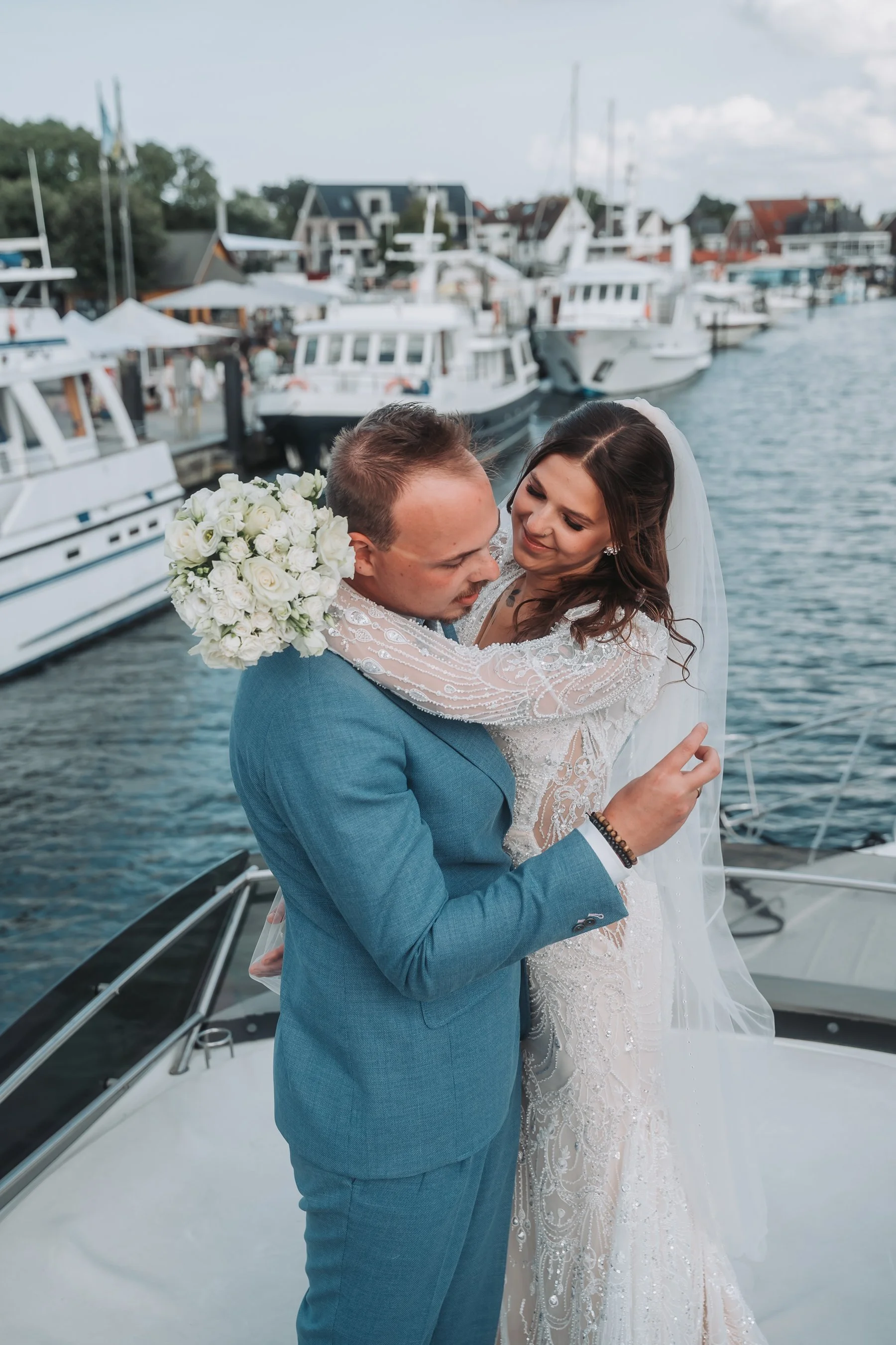 A bride and groom embrace on a boat docked at a marina, with boats and houses in the background.