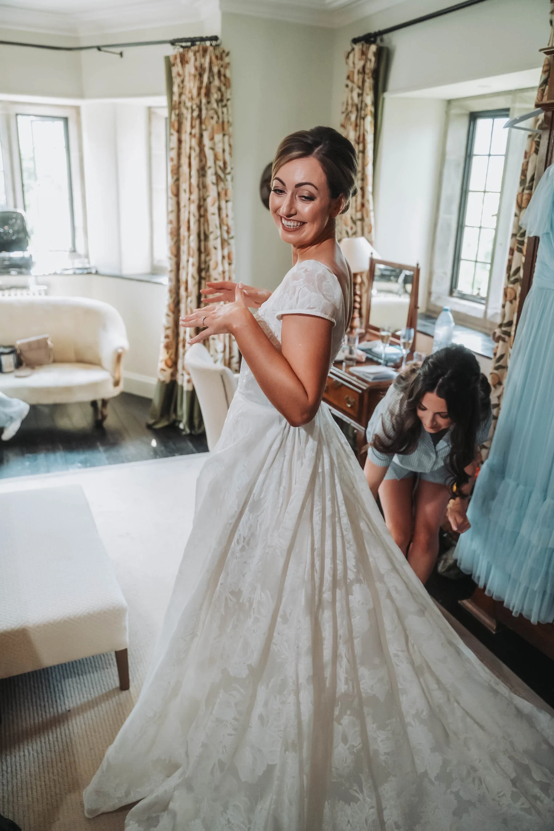 A woman in a white wedding dress smiling and turning around in a decorated room, with a second woman behind her adjusting the dress.