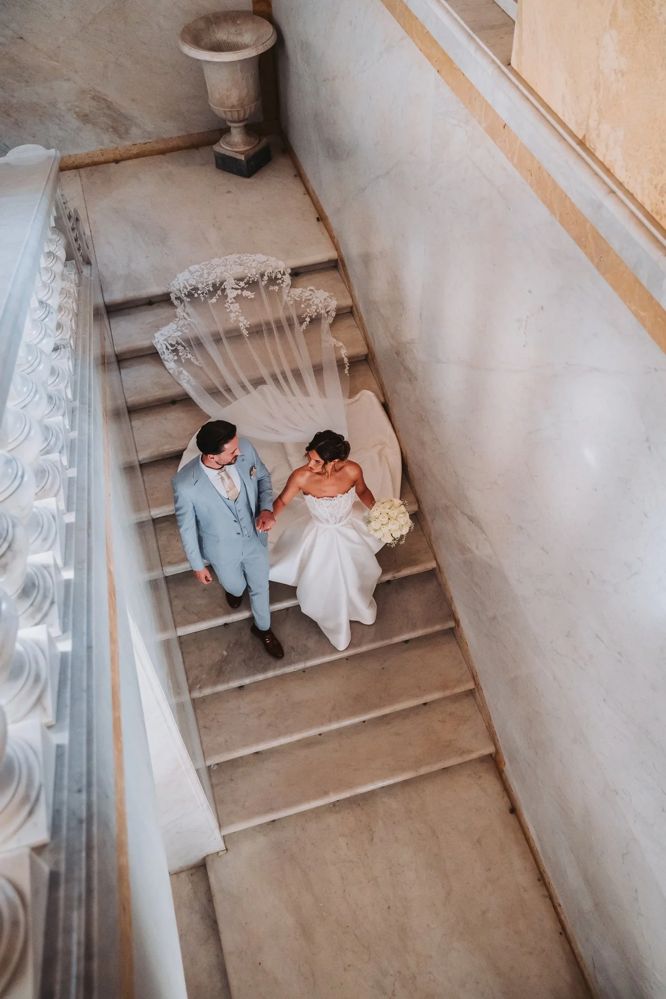 A bride and groom holding hands on a staircase with a white veil and bouquet of white roses.