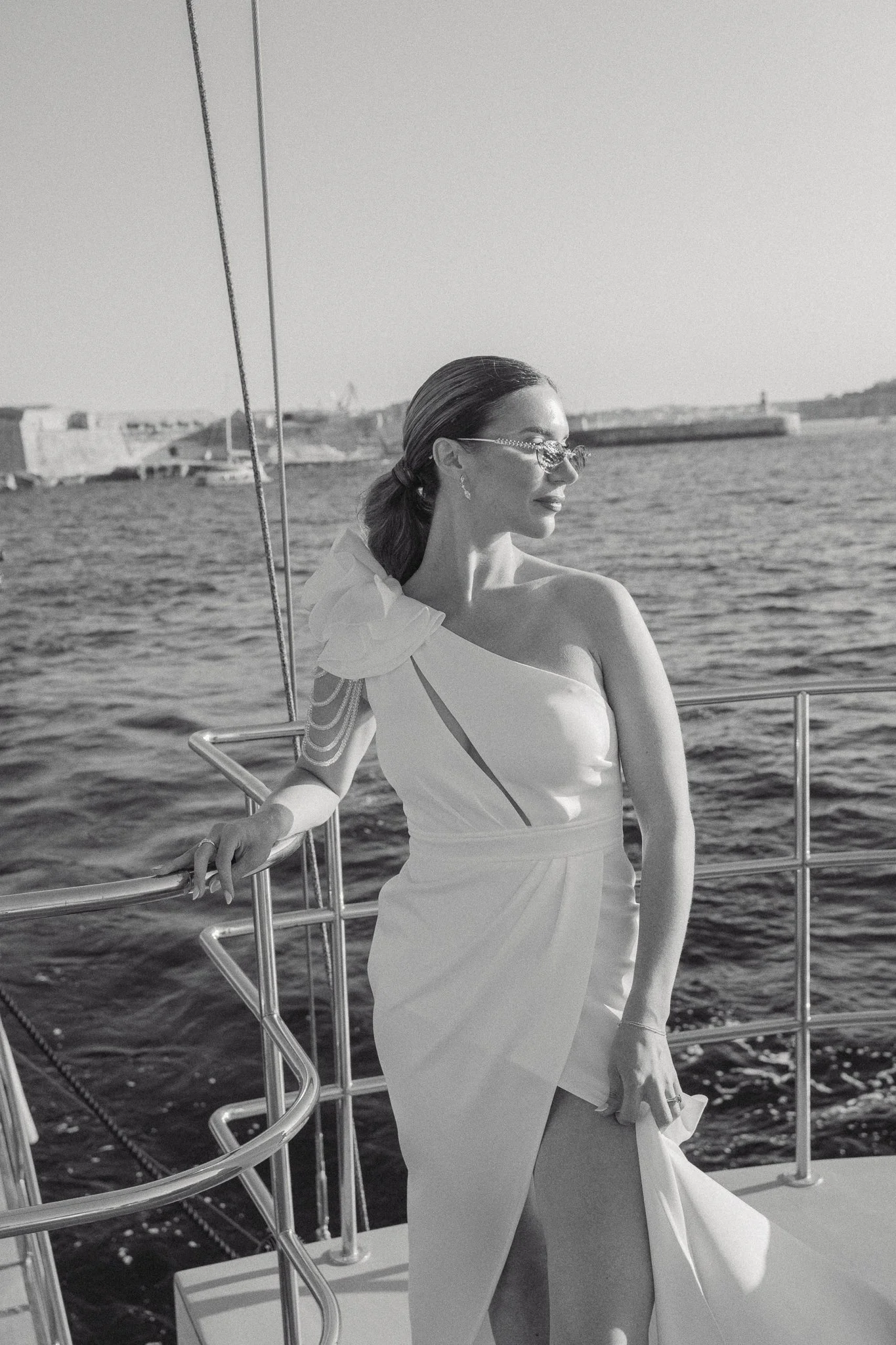 A woman in an elegant dress and earrings on a boat, holding the railing, with water and a distant shoreline in the background.