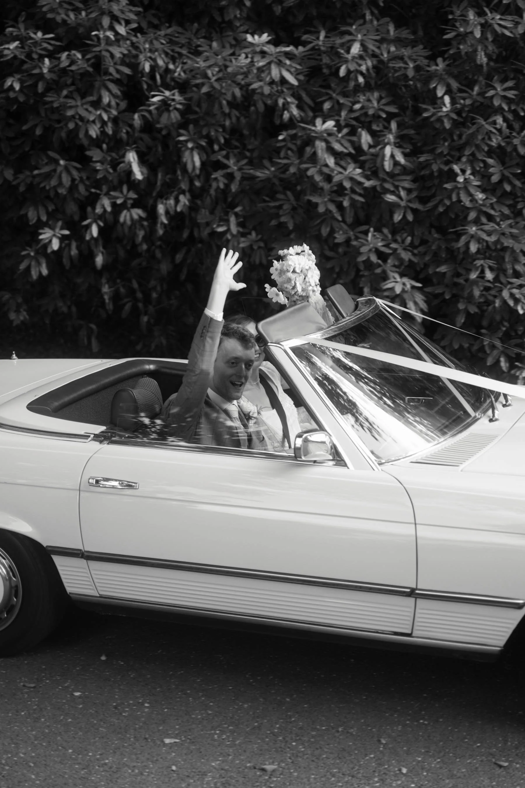A man in a suit and tie is sitting in the driver's seat of a convertible car decorated with a large floral arrangement on the dashboard. He is smiling and waving, with a large leafy tree in the background.