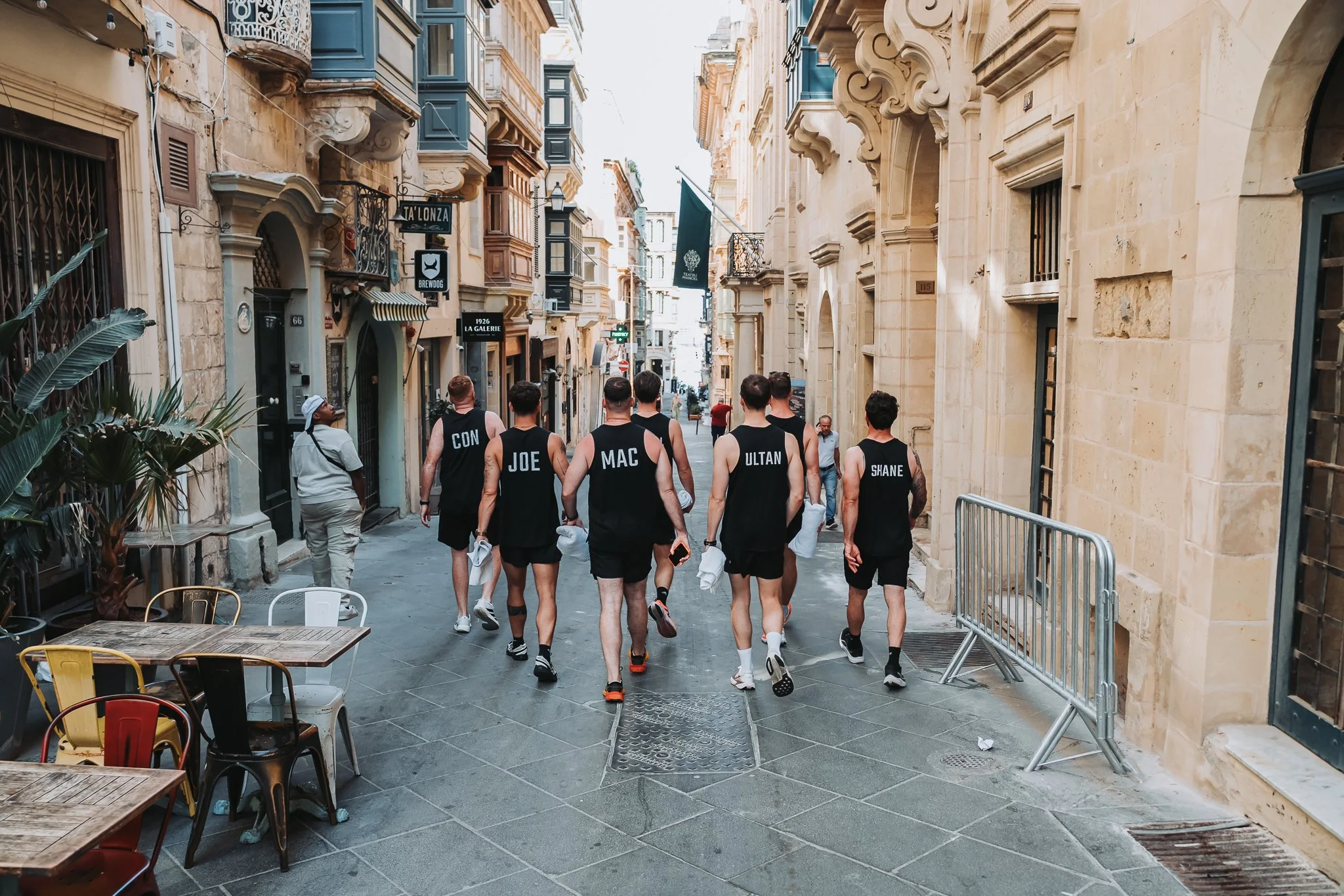 Seven men dressed in black tank tops with their names printed on the back walk down a narrow European street lined with old buildings with balconies. Outdoor tables and chairs are visible on the sidewalk.