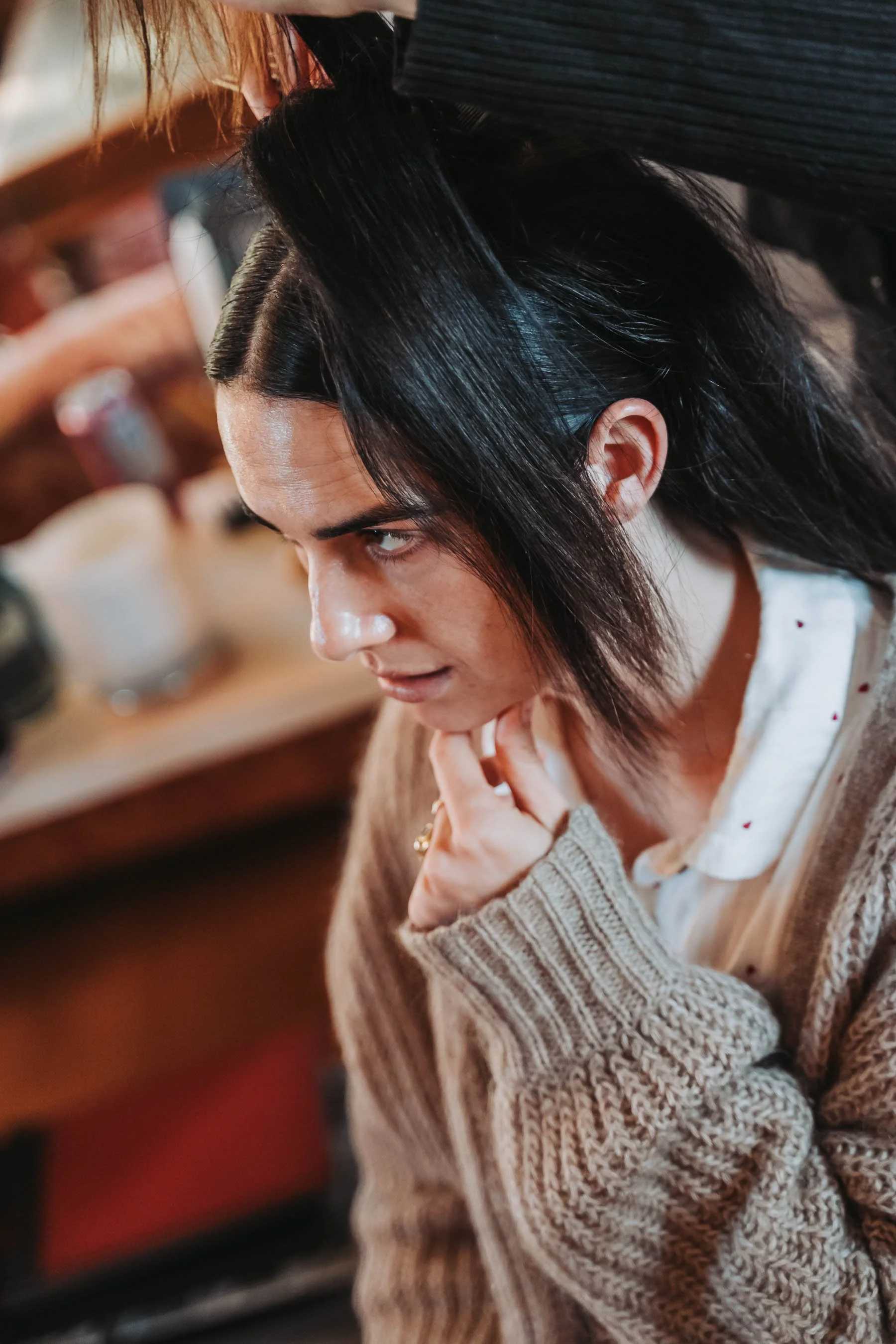 A woman with dark hair and fair skin wearing a beige knit sweater, sitting at a wooden table, resting her chin on her hand, looking thoughtfully to the side.