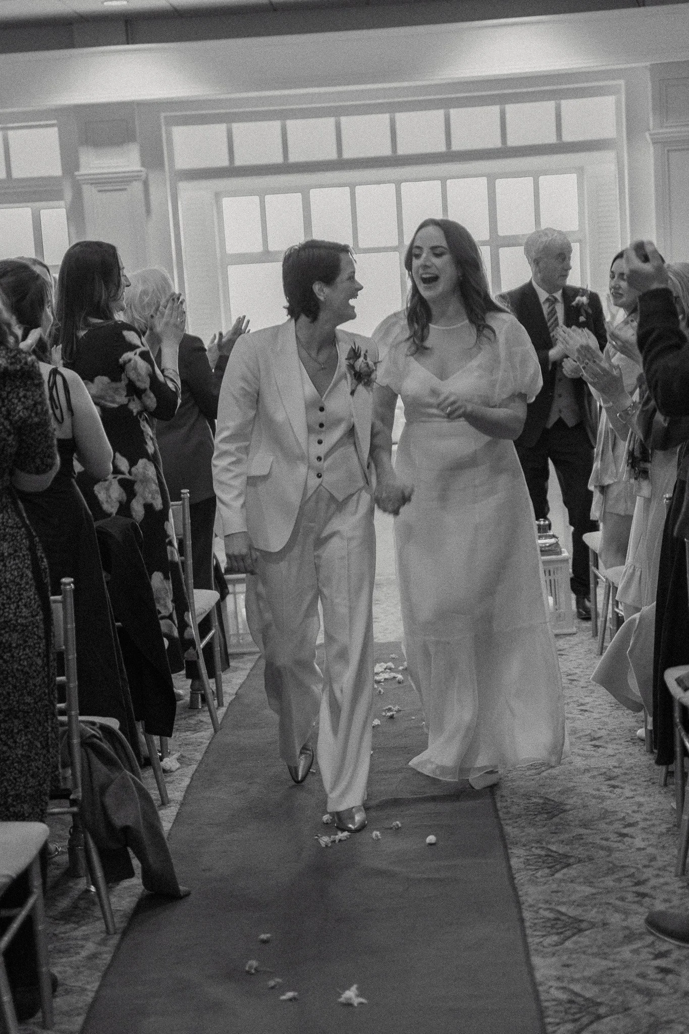 A bride and a woman, possibly her mother, walking down the aisle at a wedding ceremony. Guests are clapping and smiling on either side of the aisle, which is decorated with flower petals. Large windows in the background let in natural light.