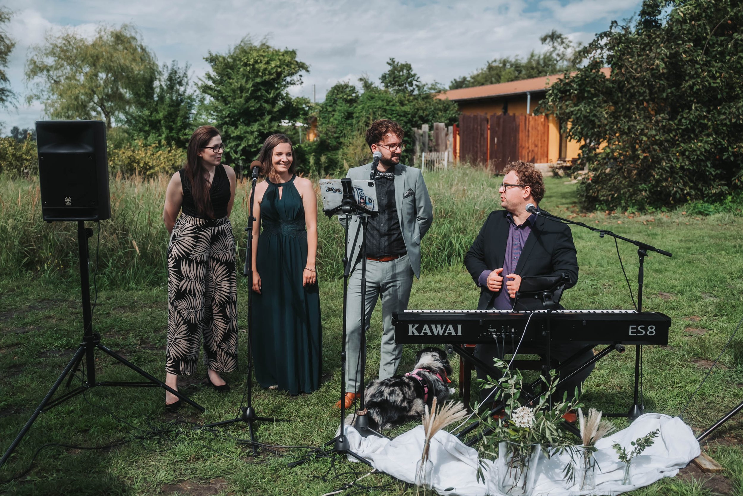 Group of five people performing music outdoors with a keyboard, microphone, and speaker, surrounded by greenery and a house in the background.