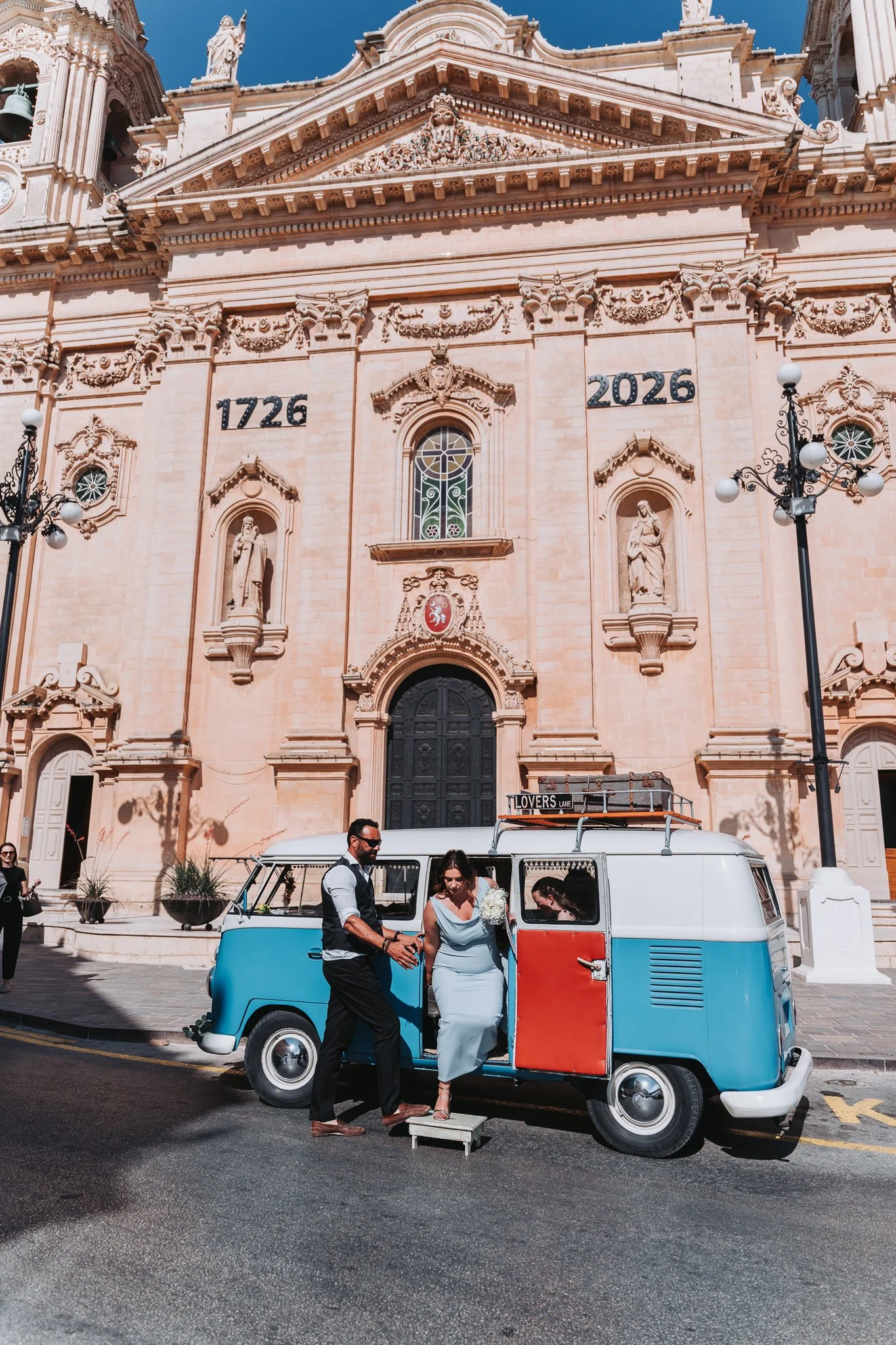 A couple in wedding attire standing beside a vintage blue and white Volkswagen van with a red door, parked in front of a historic church with elaborate architecture and statues. The setting appears to be a sunny day, and the couple is smiling at each