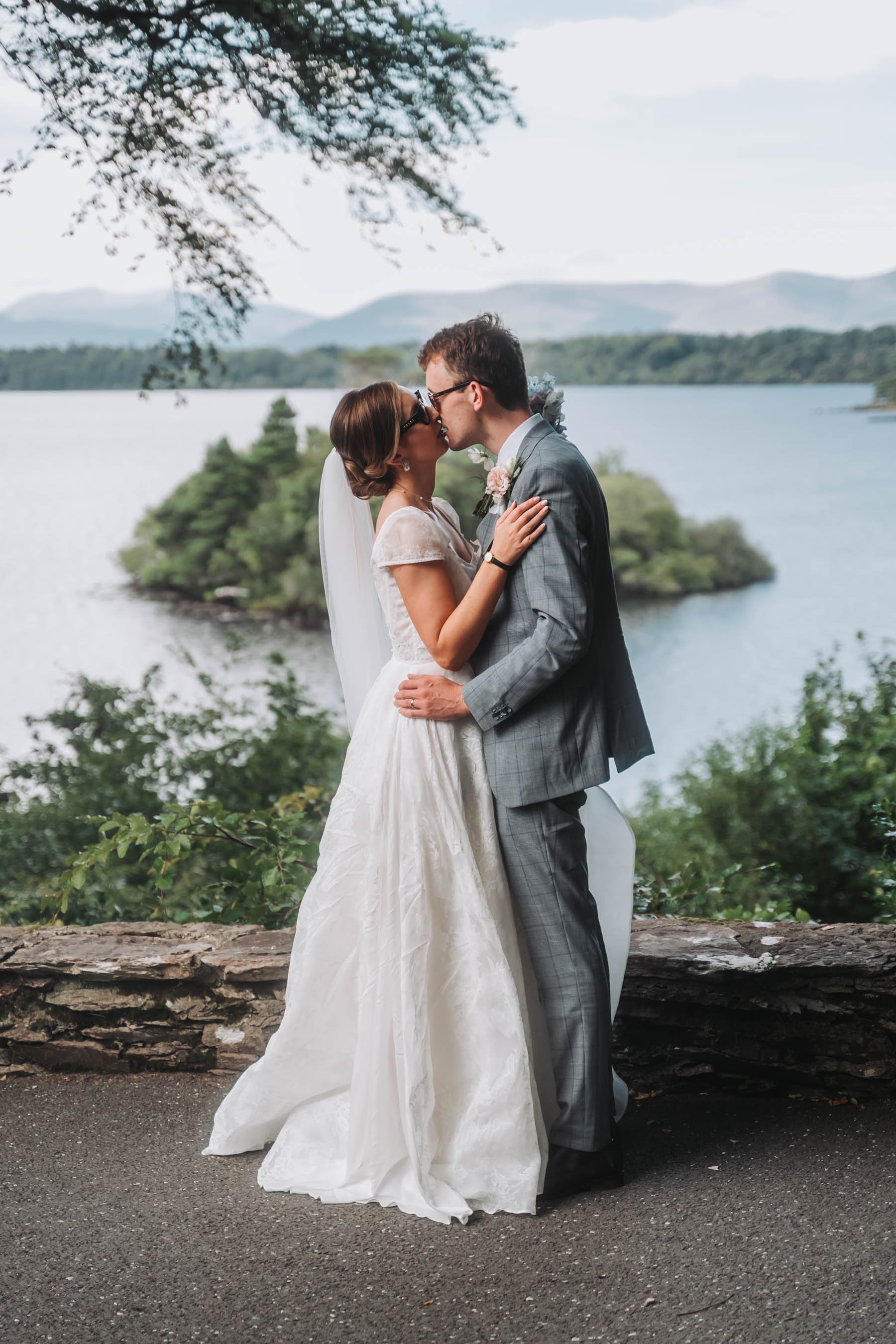 A bride and groom sharing a kiss outdoors near a lake with mountains in the background, surrounded by trees and greenery.