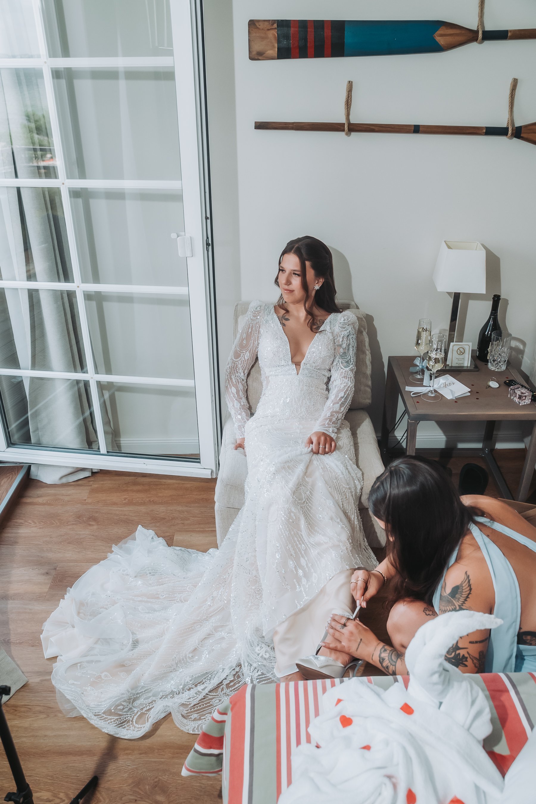 A bride in a white wedding dress with long lace sleeves sitting on a cushioned chair, while a woman with visible tattoos to the right helps her with her shoes, in a room with white walls, wooden flooring, and wall decorations resembling wooden oars.