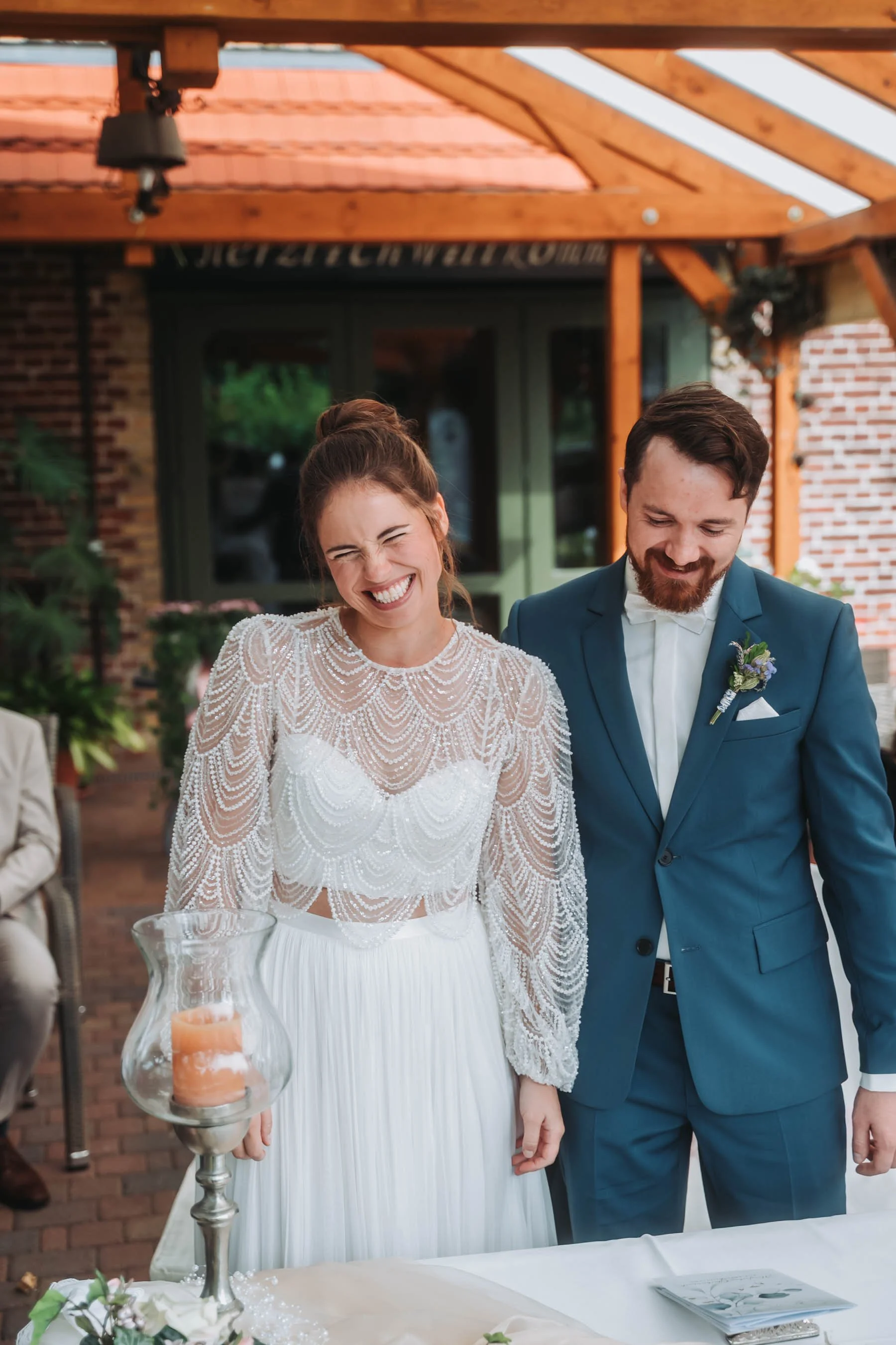 A bride and groom smiling at a wedding ceremony under a wooden pergola, with a table holding a candle holder and a bouquet of flowers in the foreground.