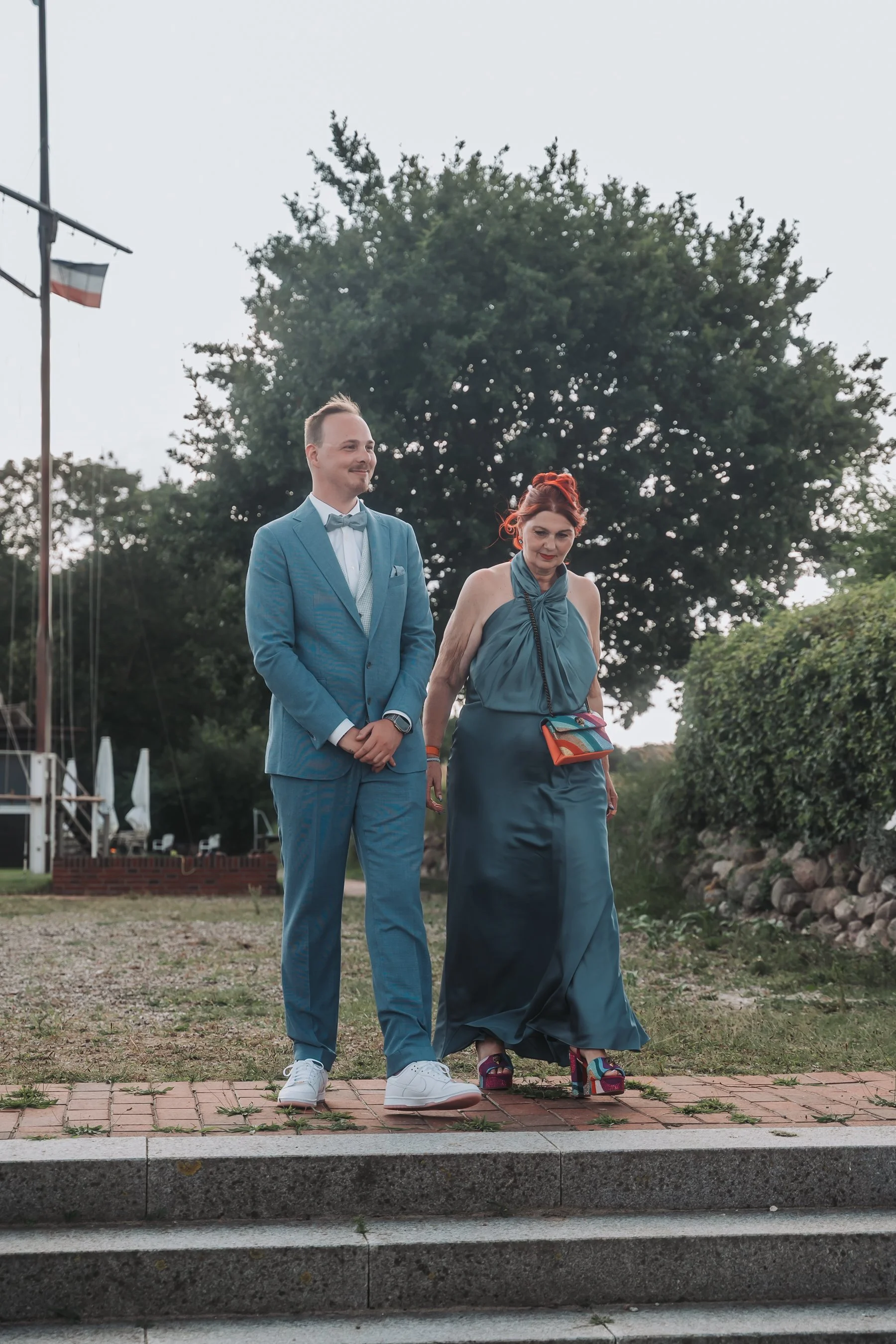 A man in a blue suit and a woman in a long blue dress walking outdoors on a brick sidewalk. The man is smiling and the woman is looking down. There are trees, a flag, and steps in the background.