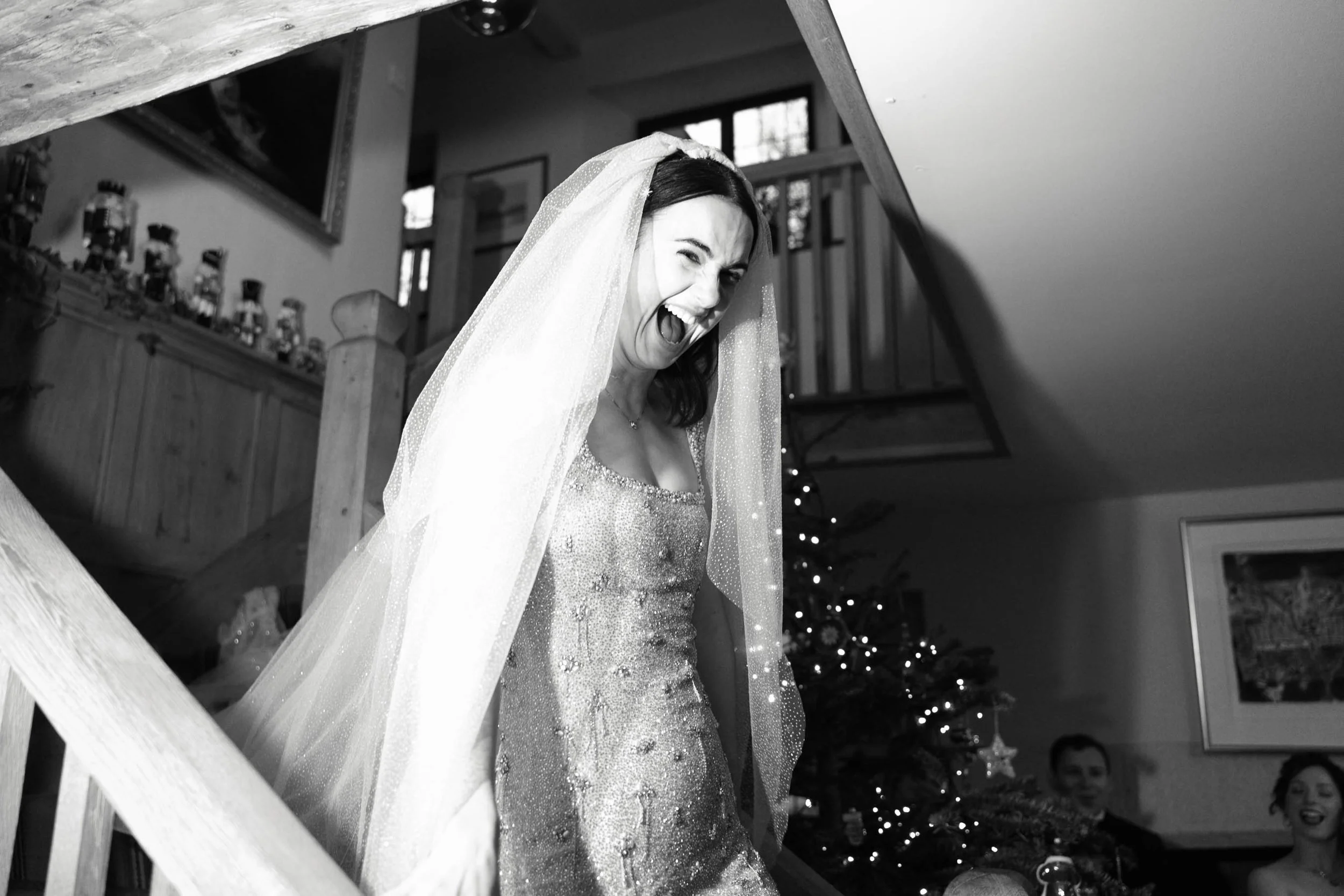 A woman with dark hair wearing a wedding dress and veil, smiling and winking. She is indoors, with a Christmas tree and other decorations visible in the background.