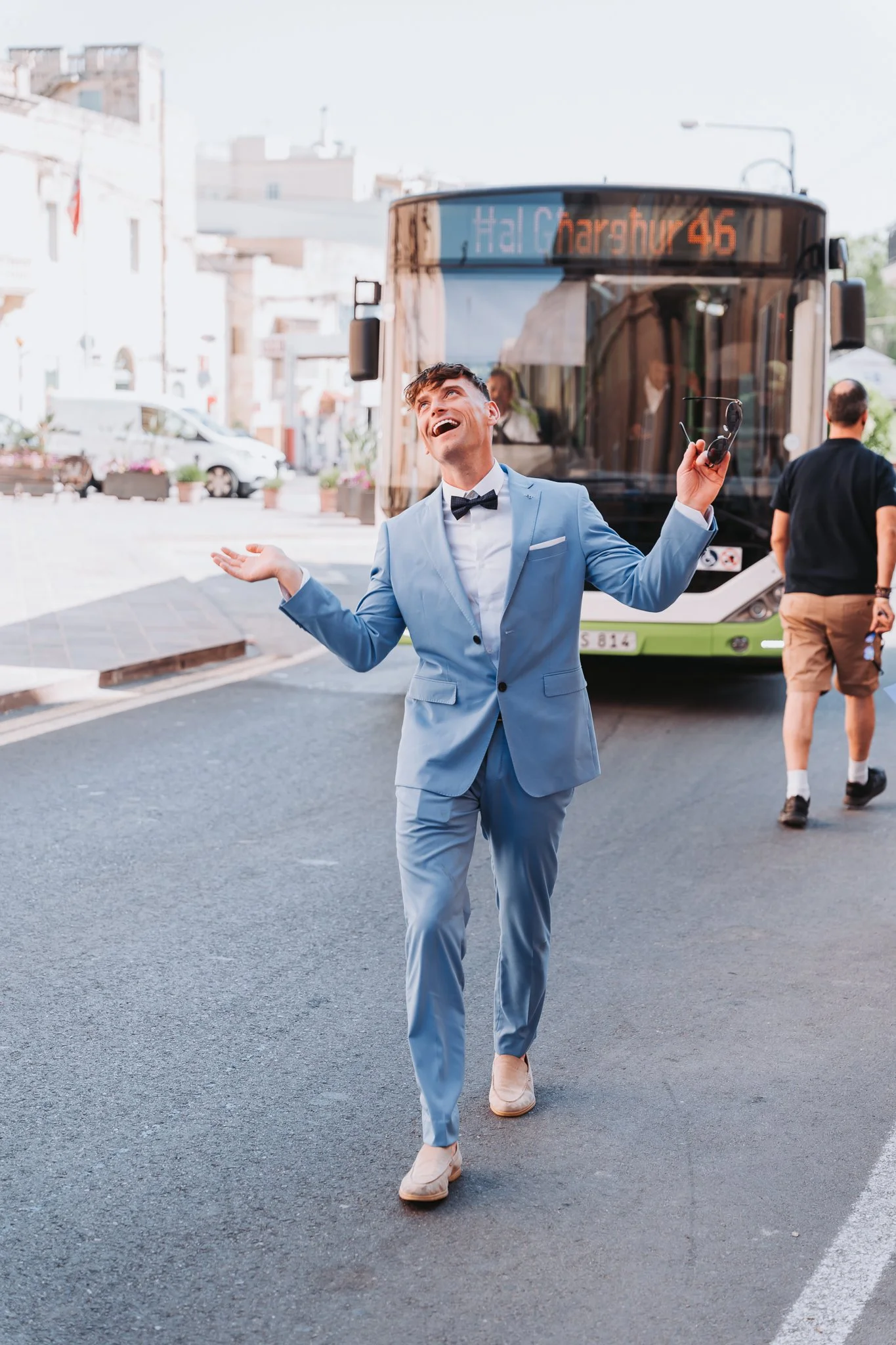 A man dressed in a light blue suit and white shirt, with a black bowtie, is smiling and walking on a city street while holding sunglasses in his right hand. A bus displaying route information is behind him, and there are other pedestrians and vehicle