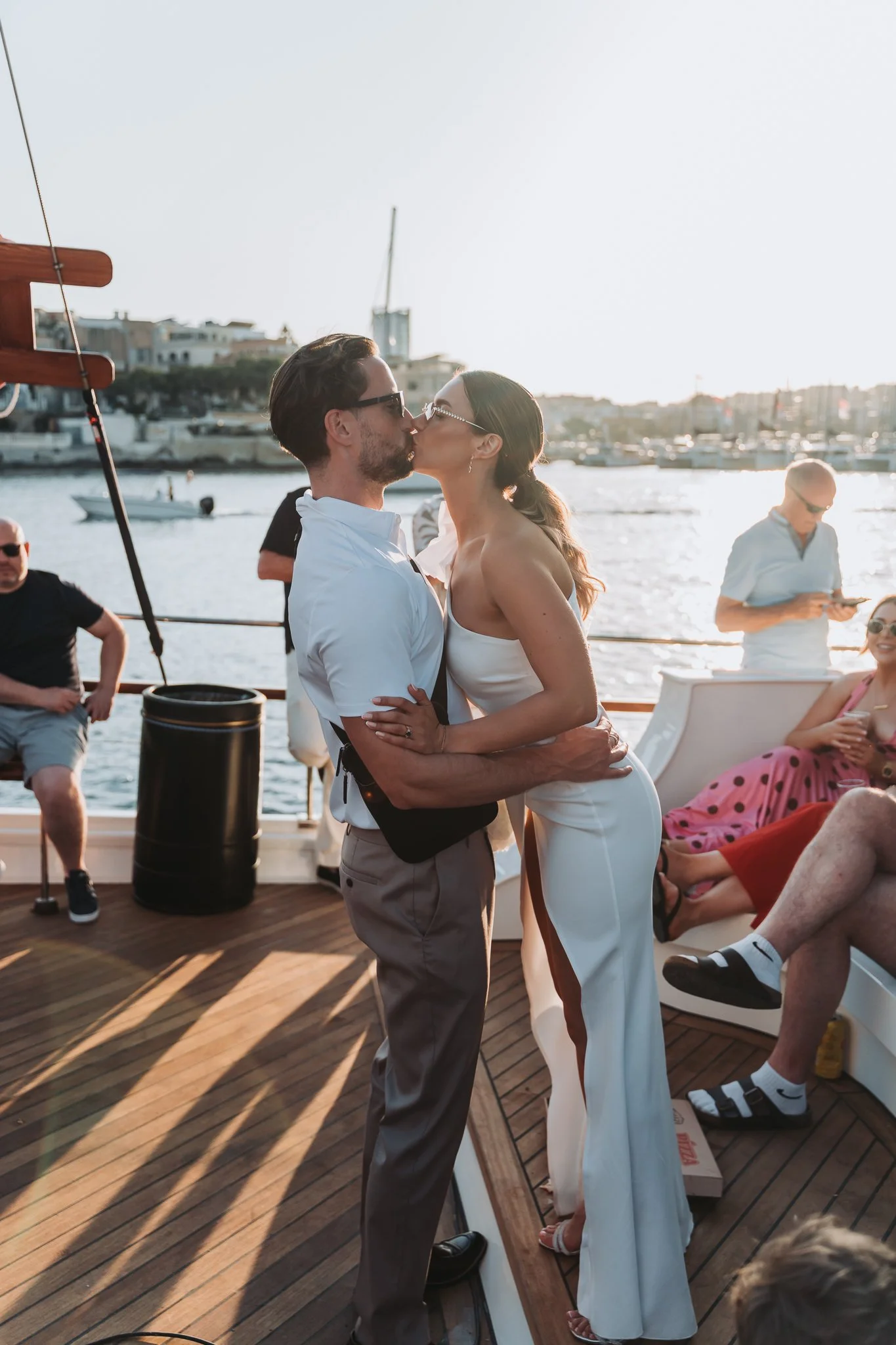 A couple kissing on a boat deck near a marina, with city buildings and boats in the background during sunset.