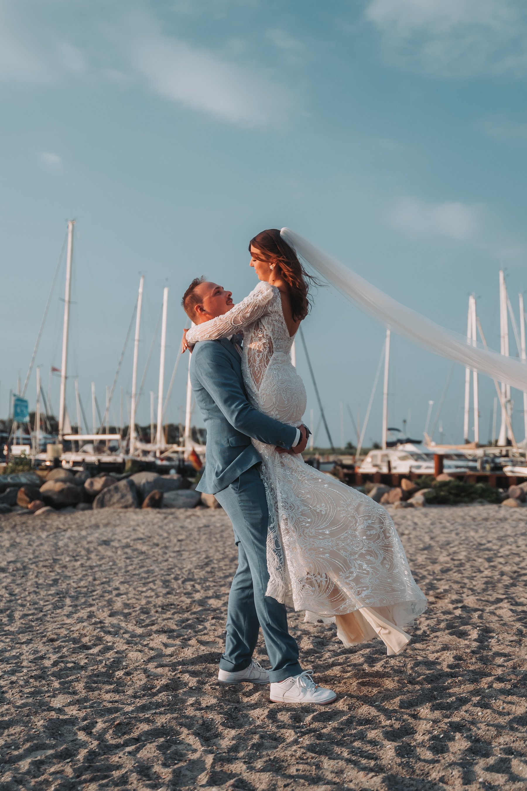 A newlywed couple dancing on a sandy beach with a marina of sailboats in the background. The groom is lifting the bride, who is wearing a lace wedding dress with a long veil, and both are smiling.