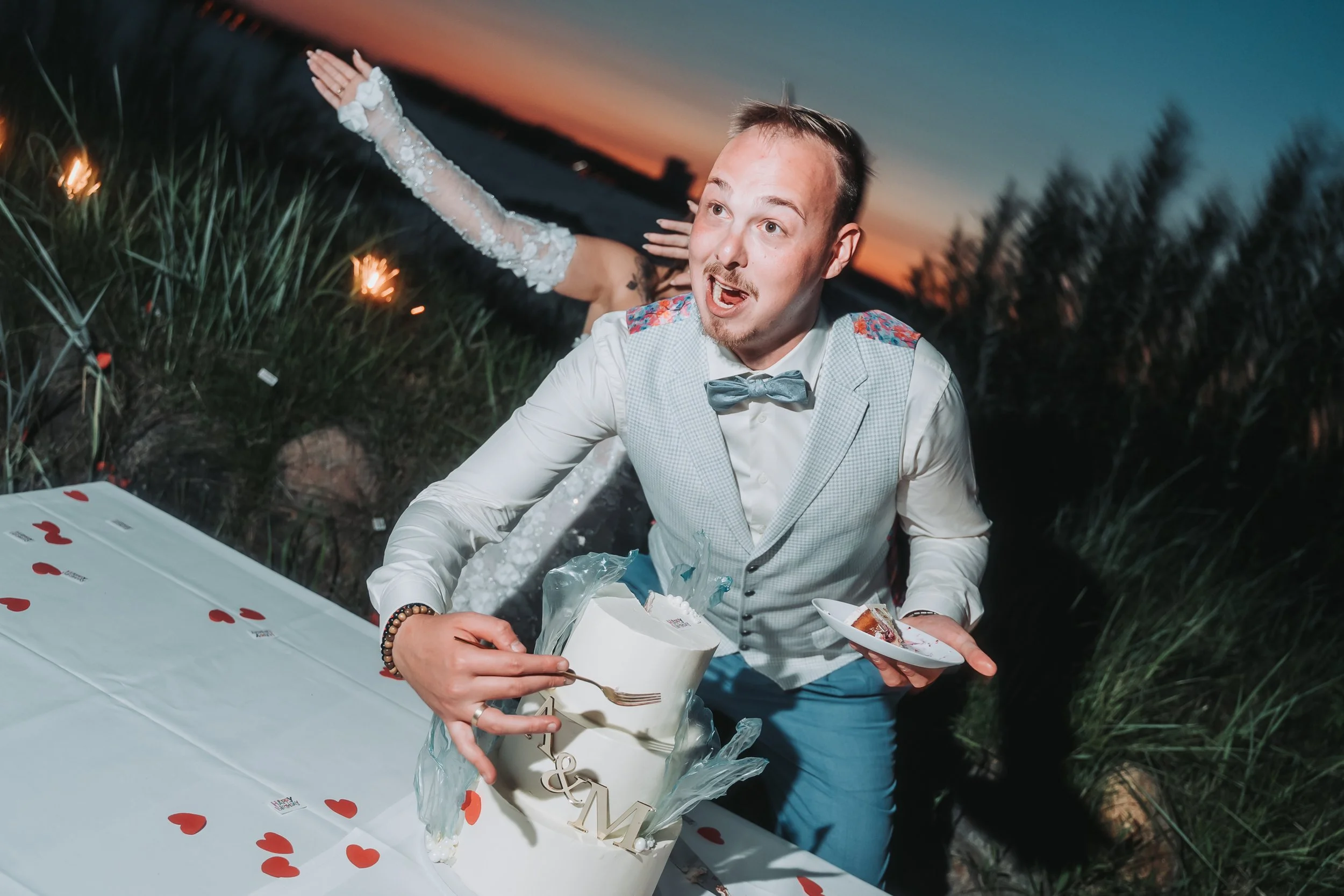 A man in a vest with a bow tie and a woman in a wedding dress celebrating outdoors at sunset, with sparklers in the background, as they reach for a cake with the word 'LOVE' on it during a wedding reception.